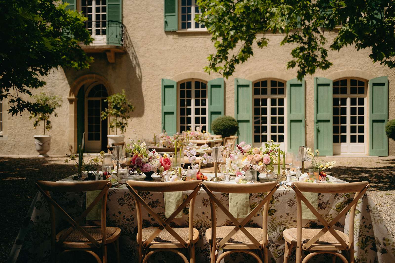 Outdoor courtyard reception tables with botanical-print cloths, pink peonies, green taper candles, and cross-back chairs