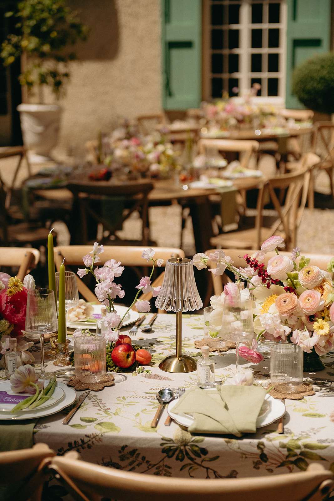 An outdoor wedding reception tablescape set in a courtyard in front of a French country house with sage green shuttered windows. Multiple round tables are dressed with a botanical-print tablecloth in cream with green foliage motifs, paired with natural wood cross-back chairs. The foreground table features a close-up detail showing a glass and brass table lamp as a centerpiece, surrounded by low floral arrangements of blush and peach ranunculus, hot pink peonies, pale pink sweet peas, yellow daisy-like blooms, and deep red accent flowers. Fresh apples and peaches are scattered among the florals as decorative elements. Place settings include white plates with sage green linen napkins, gold-toned cutlery, ribbed glassware, flower-shaped rattan coasters, small glass votive candle holders, and tall olive green taper candles. The overall styling palette is garden-inspired and romantic, mixing soft florals with fruit, botanicals, and warm metallics in an al fresco French country setting. Wide shot with shallow depth of field emphasizing the foreground table.