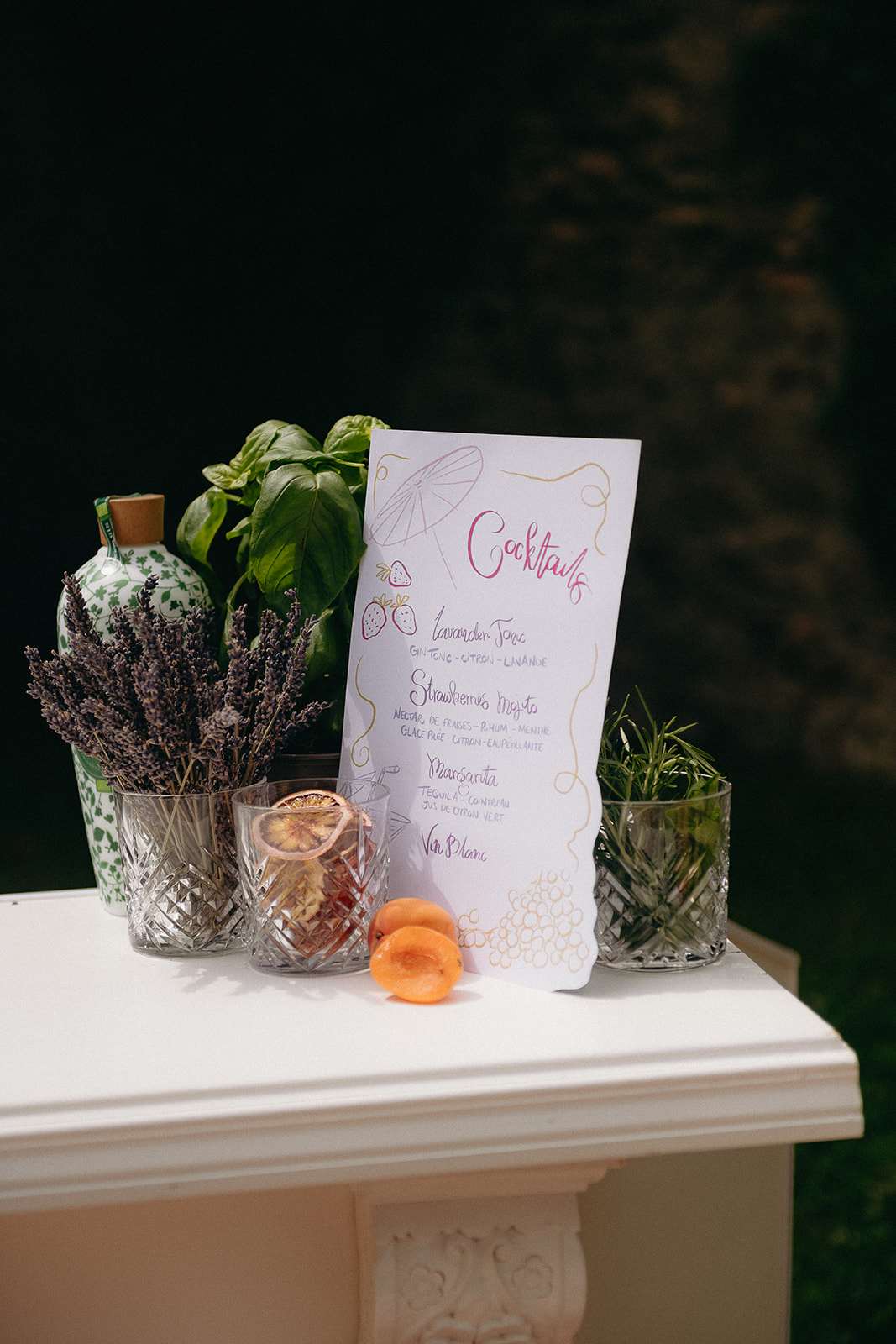 Cocktail menu in pink script with crystal glasses, dried lavender, gin bottle, and fresh herbs on plinth