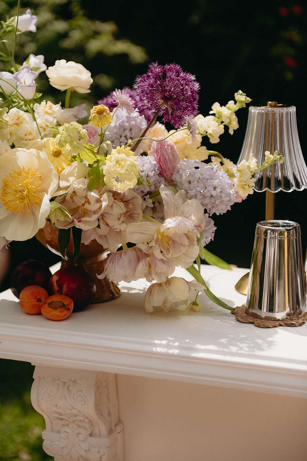 Close-up detail shot of a floral arrangement displayed on a white ornate mantelpiece or console table at an outdoor wedding, likely during a reception or cocktail hour. The arrangement is a lush, overflowing composition featuring cream and blush parrot tulips, ivory garden roses, purple alliums, lavender hyacinths, pale yellow scabiosa, small yellow daisy-like blooms, lavender sweet peas, and pink clover-style blooms, all set in a warm amber-toned vessel. Fresh apricots and dark plums are scattered to the left of the arrangement as decorative styling elements, and two scallop-edged glass and brass table lamps — one lit and one unlit — sit to the right on a woven coaster, contributing to a garden-party aesthetic with romantic, antique-inspired accessories. The overall decor palette combines soft pastels with deep purple accents and warm gold metallic details, reflecting a classic yet garden-fresh styling approach.