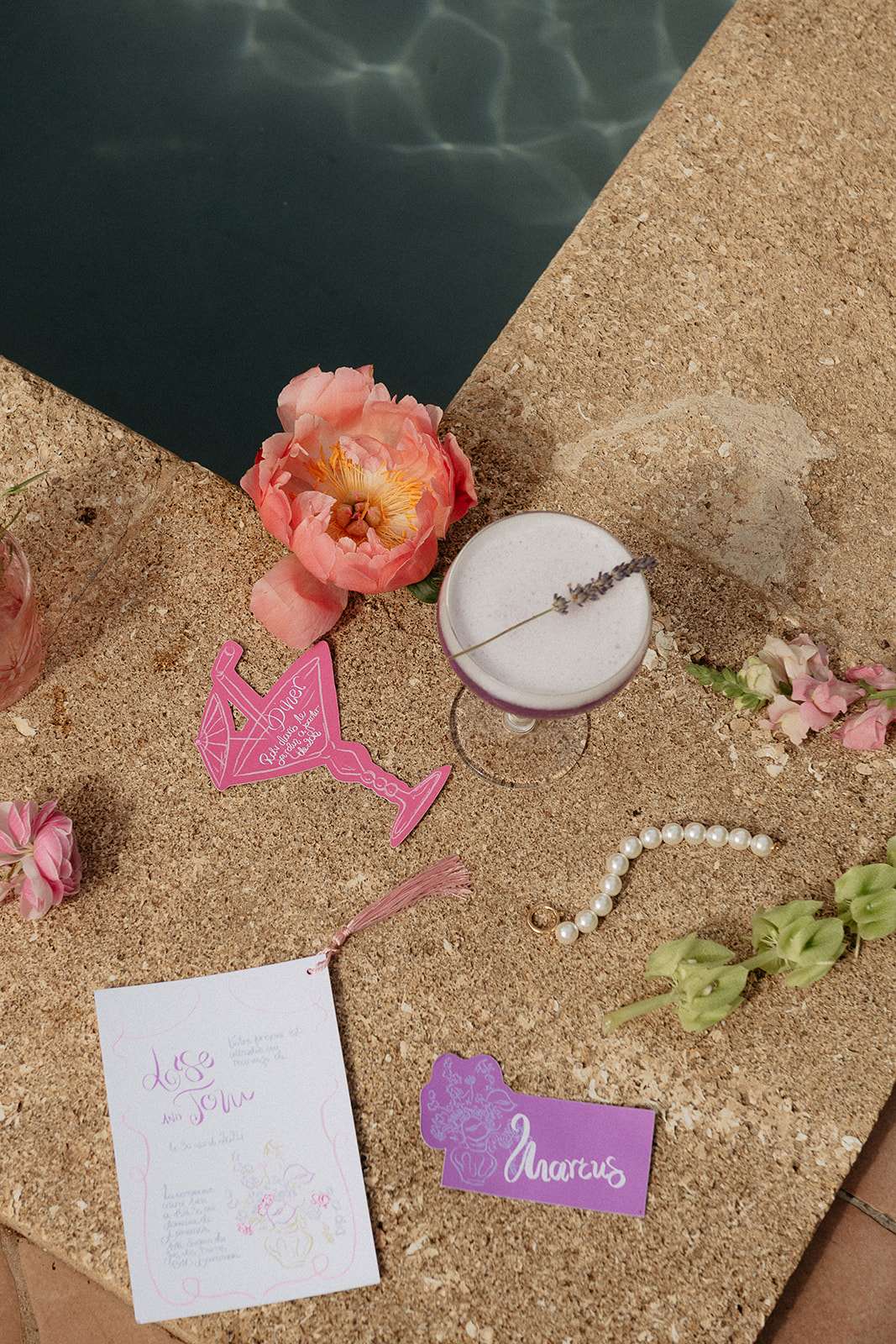 A flat lay detail shot arranged on the edge of a pool surround, with teal pool water visible in the upper left corner. The styled items include a frothy cocktail in a coupe glass garnished with a dried lavender sprig, a hot pink die-cut cocktail menu card shaped like a martini glass with a pink tassel, a white printed menu or invitation card with hand-lettered calligraphy reading 'José' and 'Tom' in lilac and pink ink, a purple place card with white calligraphy reading 'Marcus', a pearl bracelet, two gold rings, a coral-pink peony bloom, scattered small pink ranunculus and green flower buds. The overall stationery and decor palette is hot pink, lilac, and coral, with a playful, modern graphic design style.
