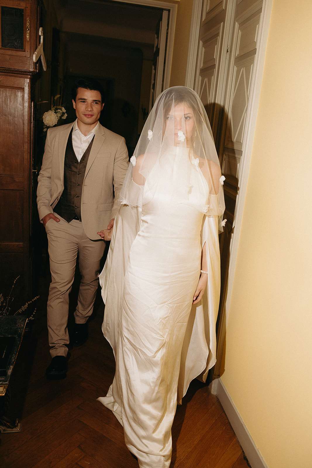 Bride in ivory satin slip gown with floral applique veil and groom in tan suit walking through chateau corridor