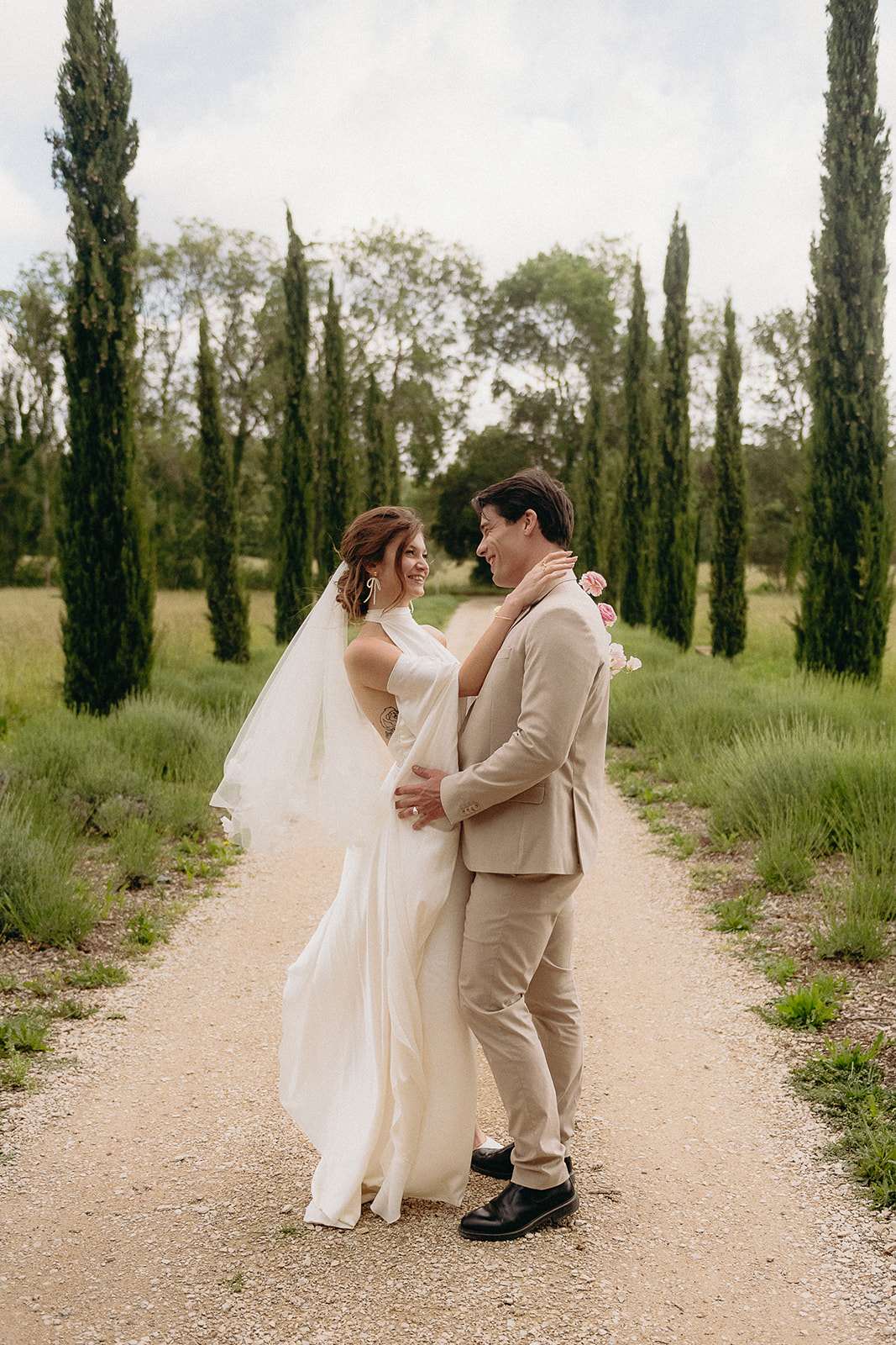 A couple portrait taken outdoors on a gravel driveway lined with tall cypress trees, suggesting a French or Mediterranean-style property. The bride wears a white halter-neck flowing gown with a cathedral-length veil, drop earrings, and has an visible back tattoo; she holds a small bouquet of blush and light pink roses. The groom wears a slim-fit sand-beige suit with black leather shoes and no tie. The two face each other closely, smiling, with the bride's hand resting on the groom's neck. The composition is a full-length portrait shot with the cypress-lined path receding into the background, giving the image depth.