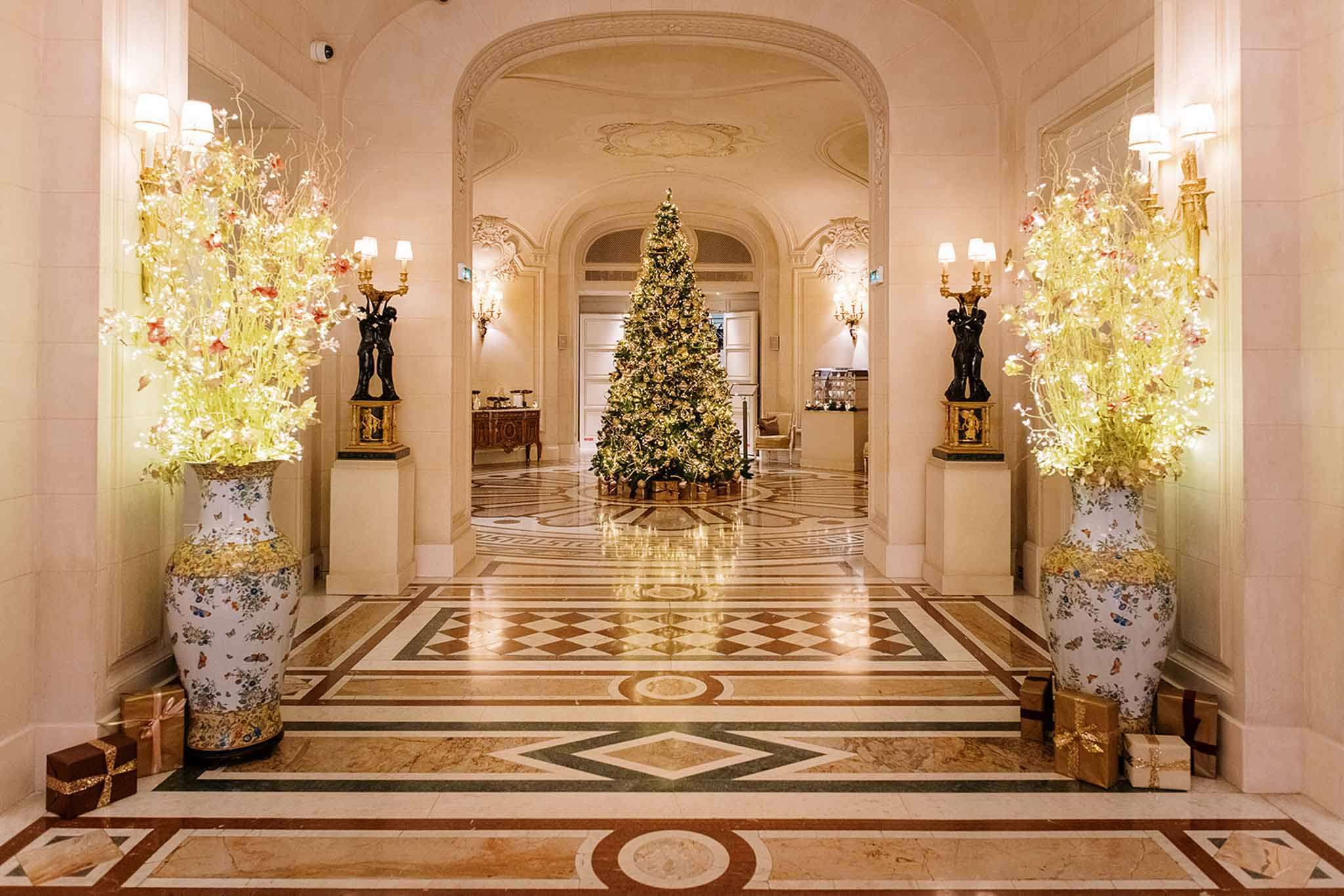 A wide shot of an indoor grand hotel or palace lobby decorated for Christmas, with no people present. The hallway features an ornate inlaid marble floor in cream, green, and brown geometric patterns, arched doorways with decorative plasterwork ceilings, and warm wall sconce lighting. Two large decorative porcelain vases with floral botanical motifs flank the entrance, each filled with tall arrangements of chartreuse and pale pink branches with fairy lights woven through. A fully decorated Christmas tree with warm white lights and gold ornaments stands at the far end of the corridor, surrounded by wrapped gifts in gold and brown at its base. Additional wrapped gifts in pink and gold are placed near the foreground vases, and pairs of bronze figural candelabras on stone pedestals line the corridor. The overall decor palette is gold, cream, and warm white, with a classic French palatial interior style. Potential venue feature image.