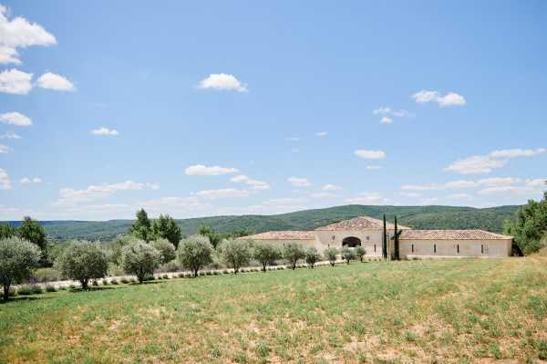 Single-storey stone venue with terracotta roof, arched entrance, olive tree row, and wooded hills beyond