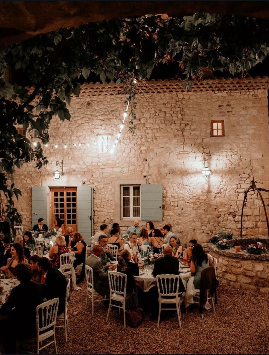 Evening courtyard reception at stone farmhouse with festoon lights, round tables, and white chairs on gravel