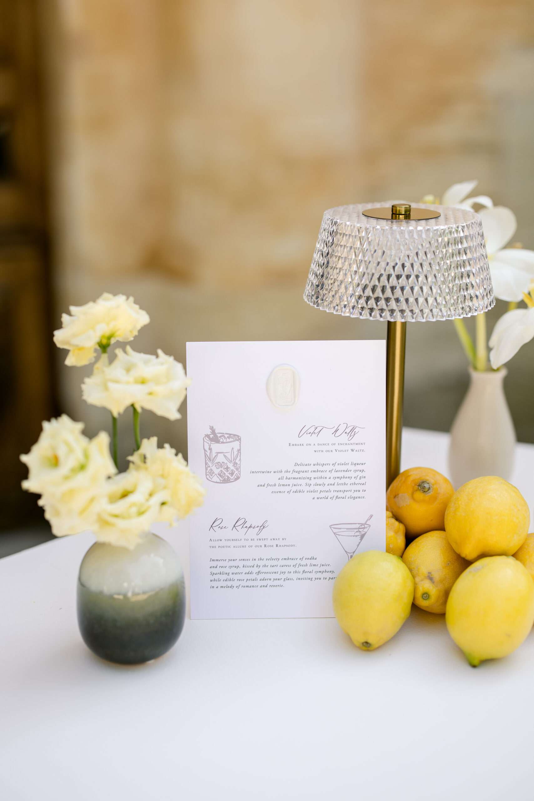 Cocktail menu card with wax seal beside crystal lamp, lisianthus, and fresh lemons on white linen