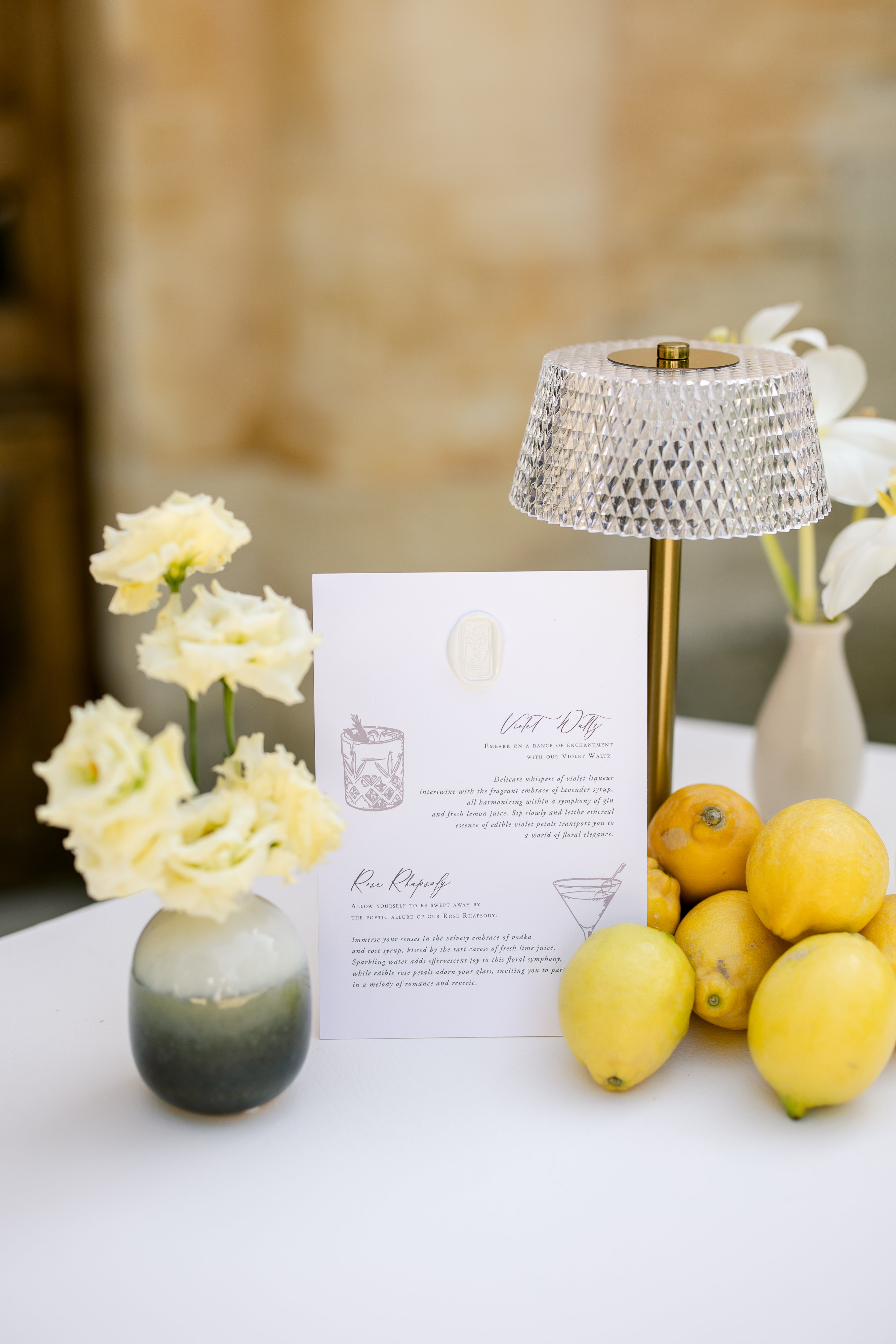 Cocktail menu with gold wax seal listing signature drinks crystal lamp cream lisianthus and fresh lemons on white surface