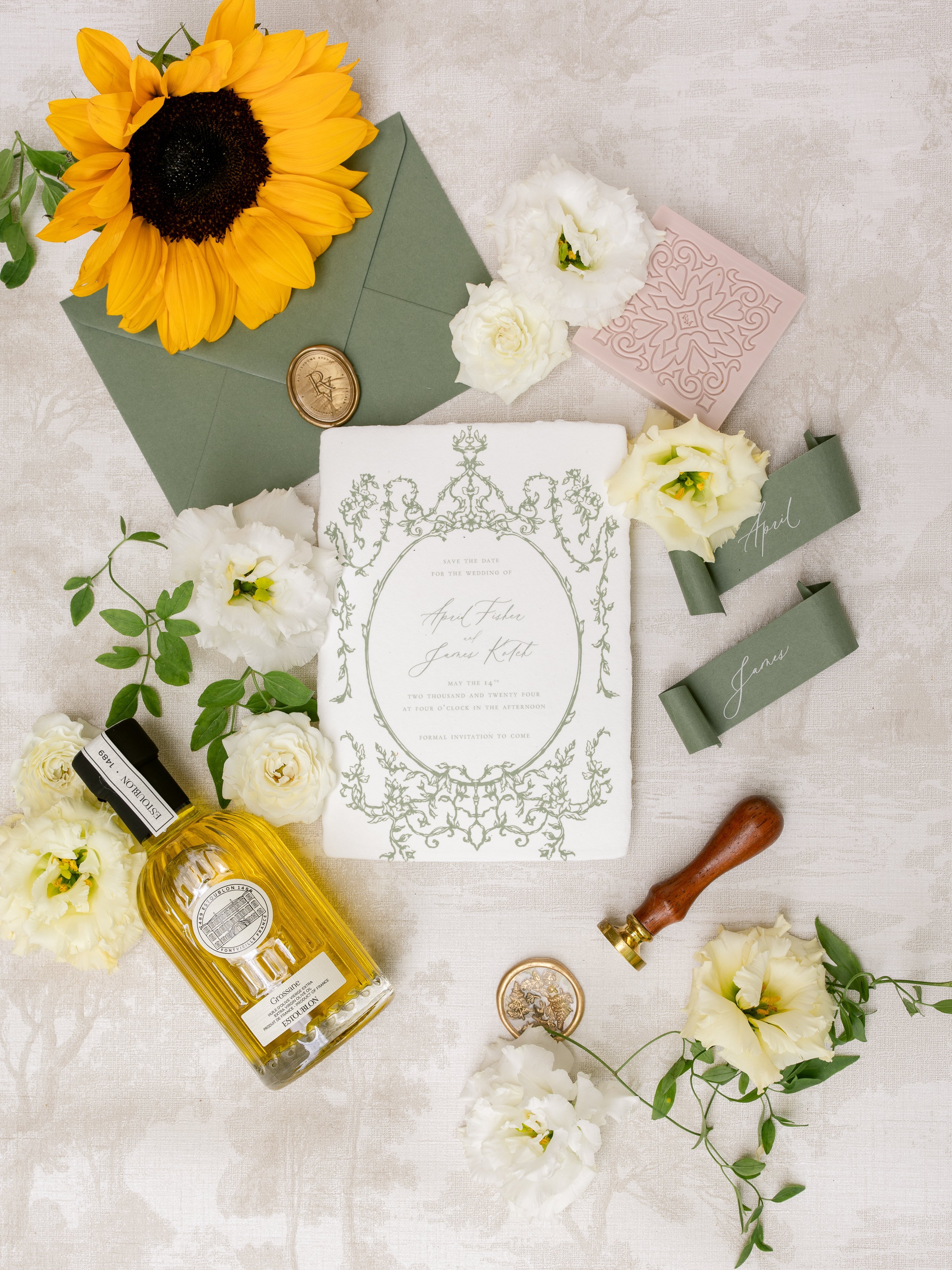 A flat lay detail shot of a wedding stationery suite arranged on a light linen-textured surface. The centerpiece is a white deckle-edge save-the-date card printed with a sage green ornate botanical frame, featuring calligraphy-style script reading 'April Fisher and James Kotel, May the 14th, Two Thousand and Twenty-Four.' Surrounding the card are a sage green envelope sealed with a gold monogram wax seal, two sage green silk ribbon place card tags hand-lettered with the names 'April' and 'James,' a blush pink embossed decorative tile or card holder, and a wooden-handled wax seal stamp with a brass head. Additional styling props include a bottle of Estourlon olive oil or fragrance with a golden-amber liquid, scattered cream garden roses, white lisianthus blooms, and a single yellow sunflower, with trailing green foliage throughout. The overall color palette is sage green, cream, and gold with a classic, French-inspired aesthetic.