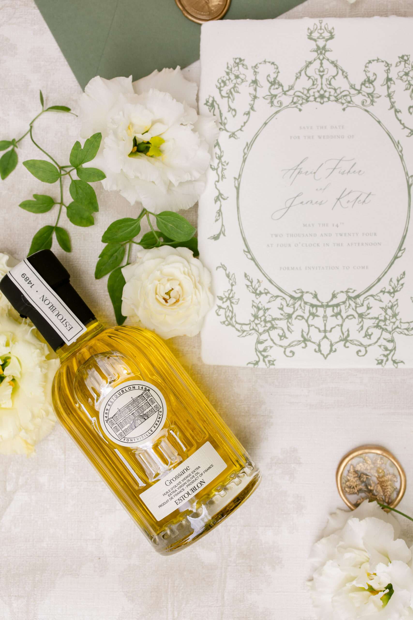 Flat lay of save-the-date card with sage green baroque frame, white roses, olive oil bottle, and gold wax seal on cream linen