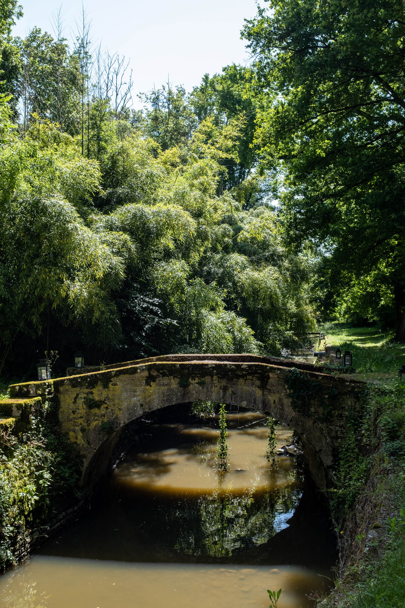 Historic moss-covered stone arch bridge spanning a shallow waterway on venue grounds with lanterns along the path