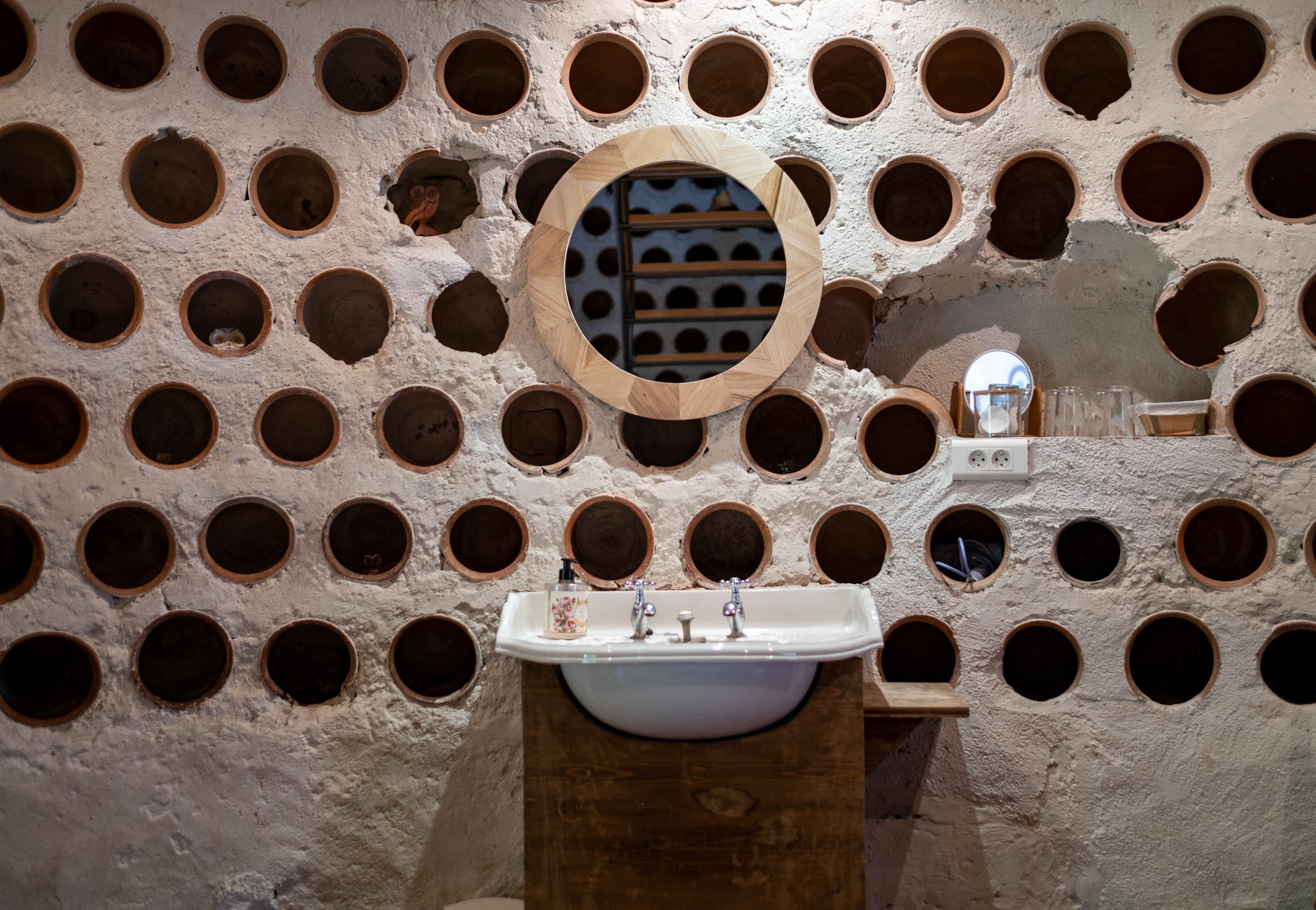 Venue bathroom with terracotta wine bottle cavity wall, white basin on raw wood vanity, and geometric wood-framed mirror