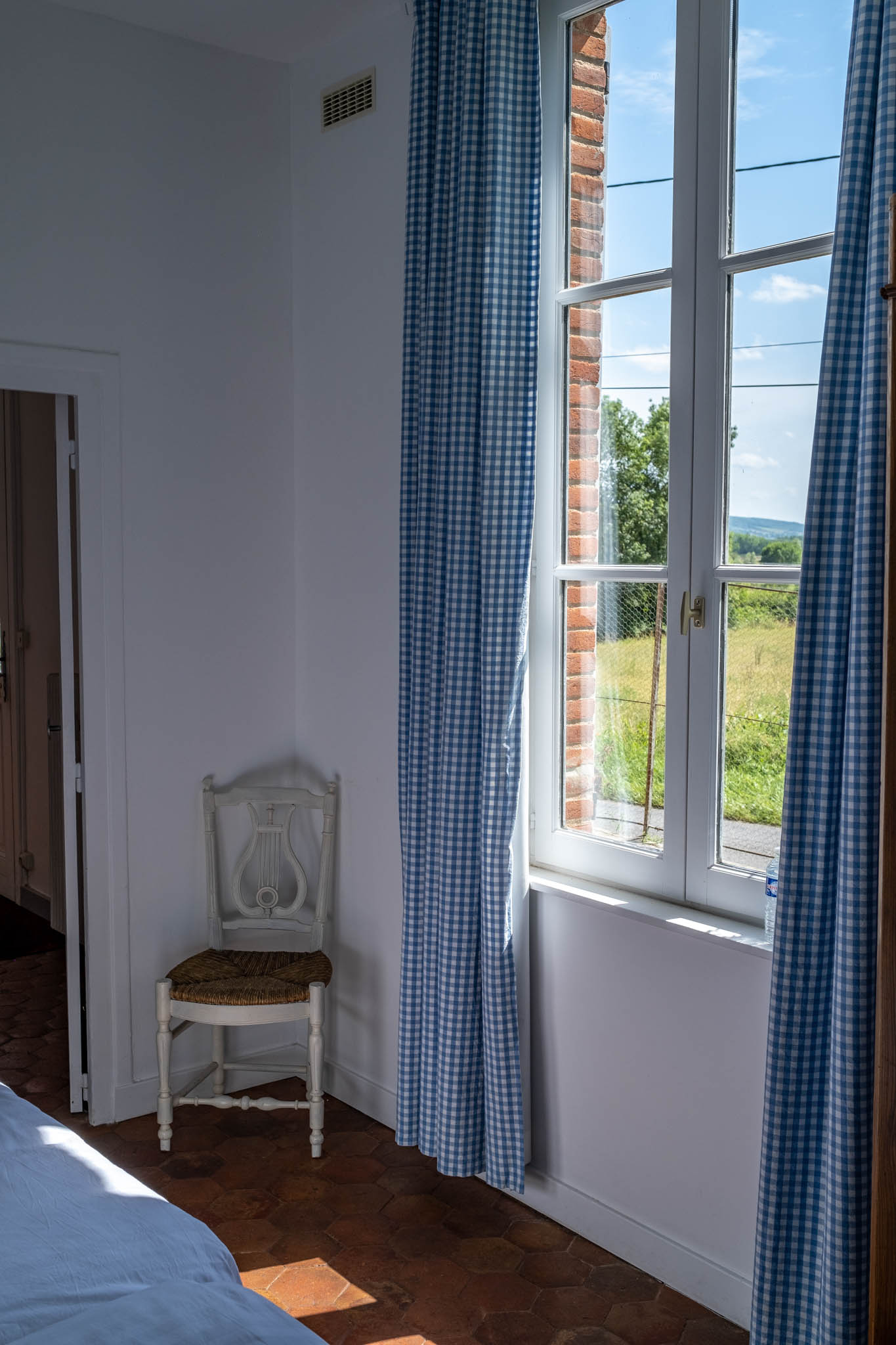 French country bedroom with white antique lyre-back chair, blue gingham curtains, and terracotta floor
