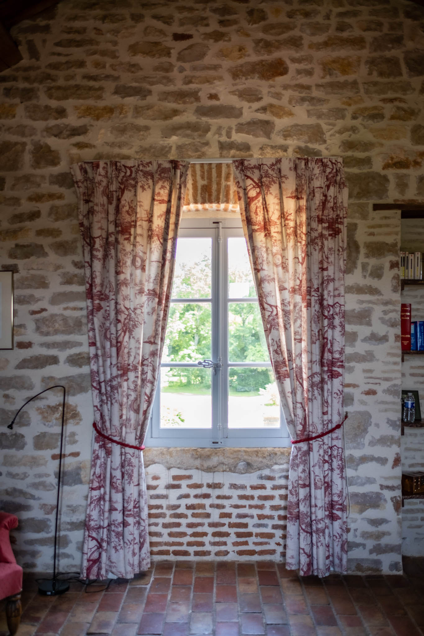 French stone room interior with toile de Jouy curtains, terracotta floor tiles, and casement window overlooking grounds