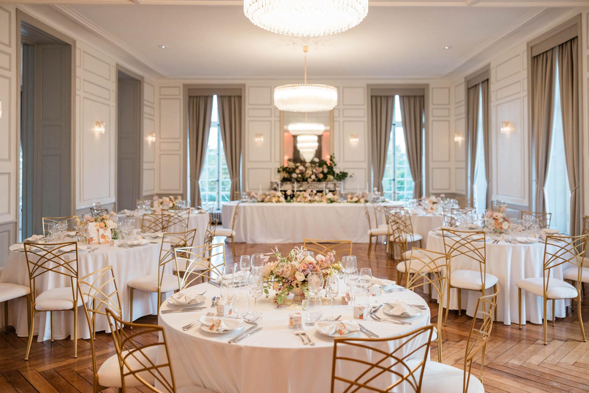Ballroom with round tables, blush rose centrepieces, gold geometric chairs, and crystal chandeliers