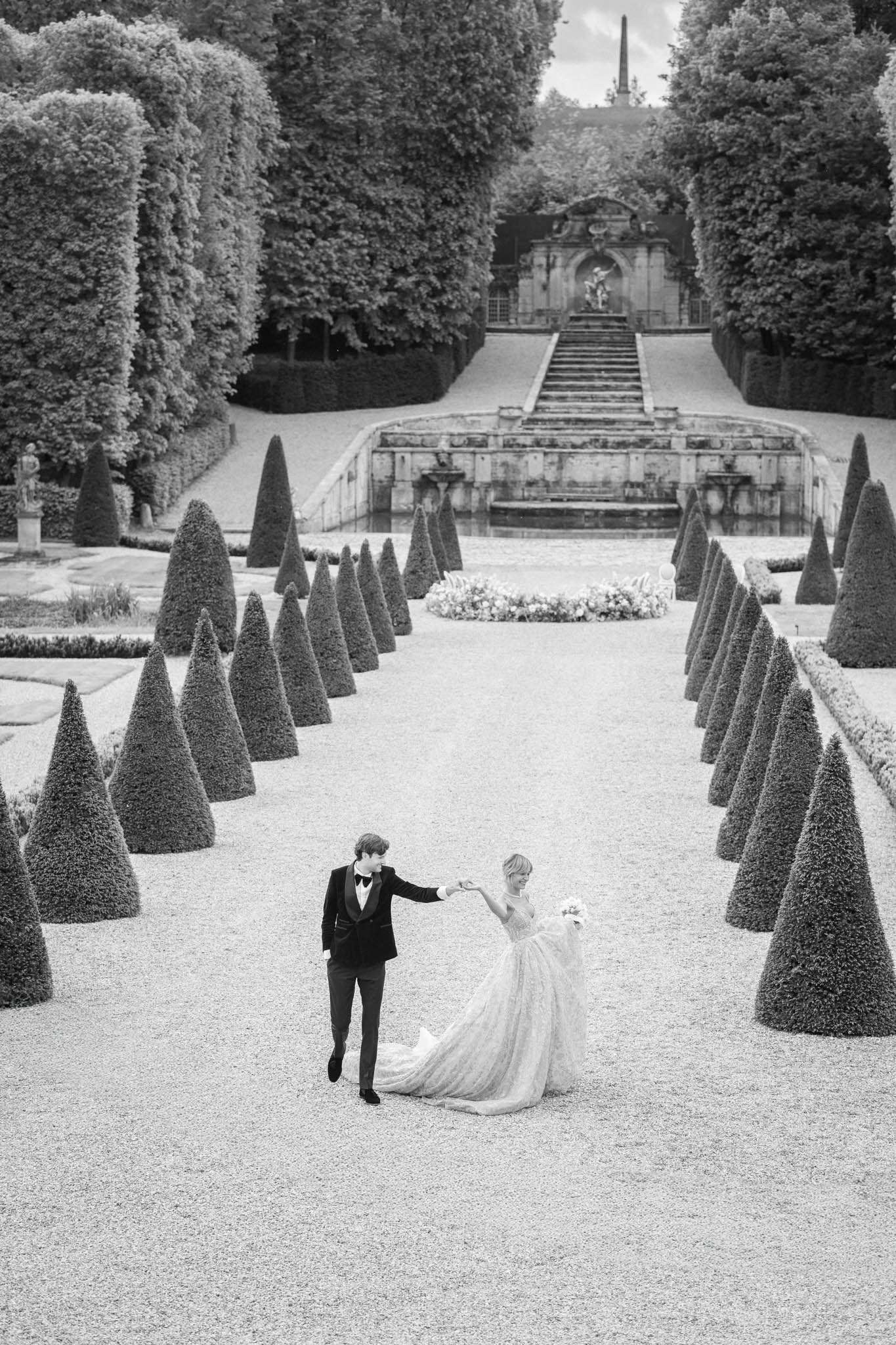 Black and white aerial view of bride and groom walking along a formal French garden allee with topiary hedges