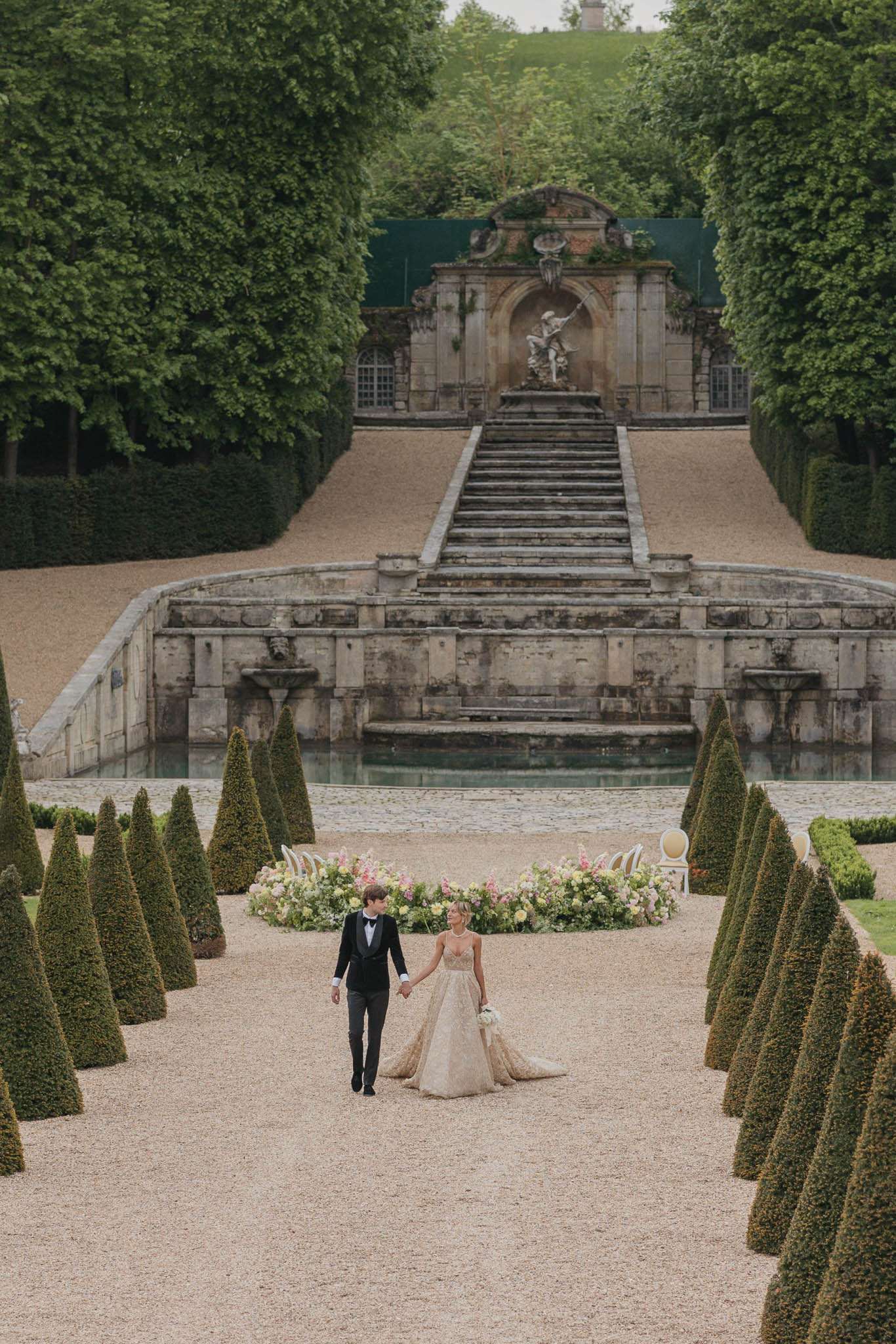 Couple walking hand in hand along formal French garden allee with topiary hedges and baroque stone staircase