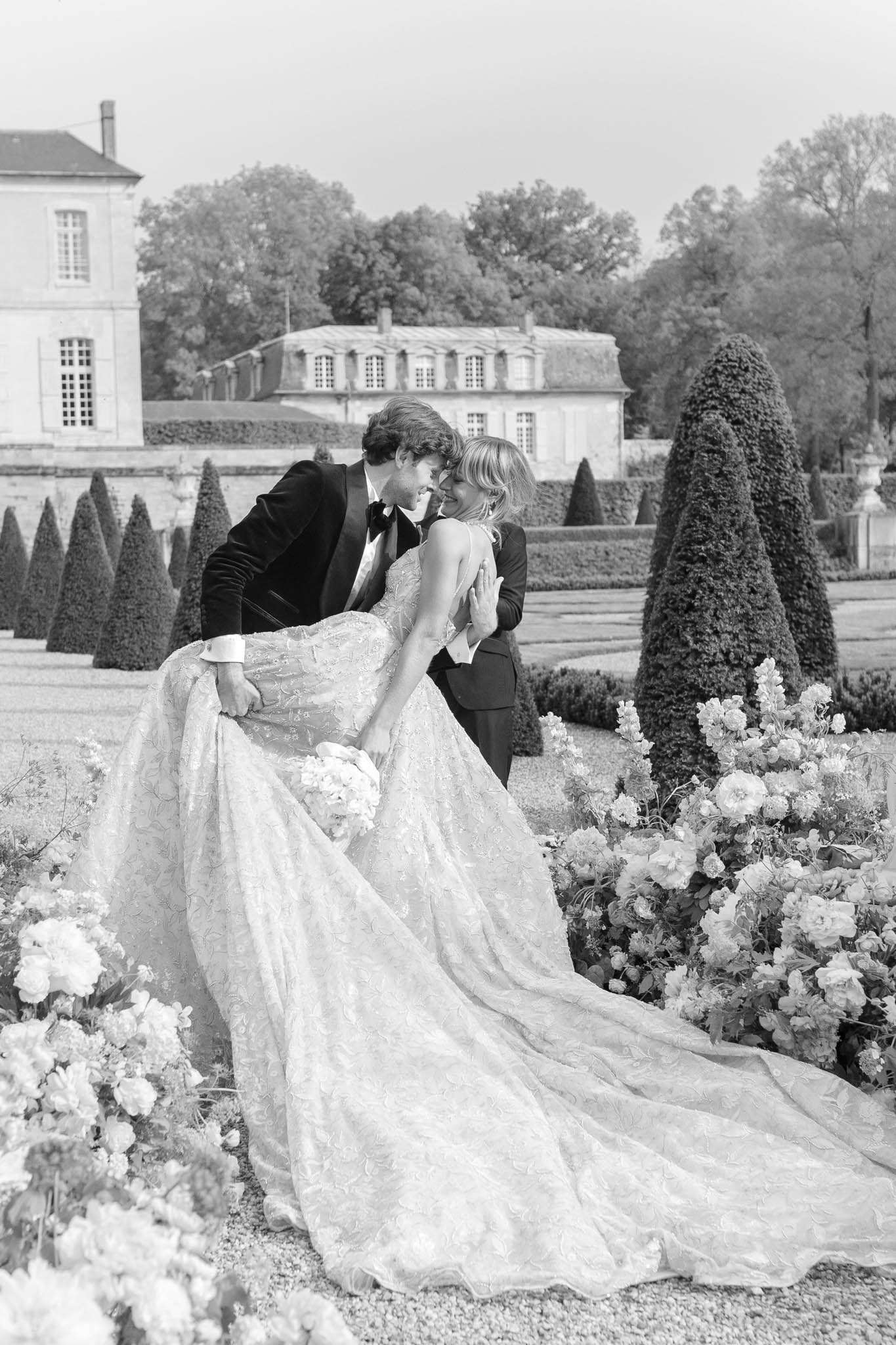 Groom lifts bride's cathedral train in formal topiary garden before mansard-roofed chateau in B&W
