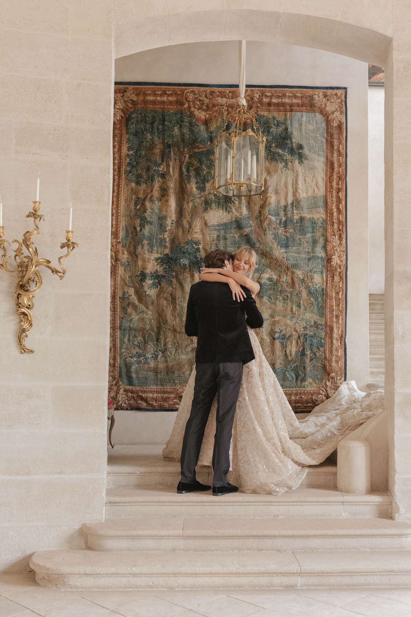 A couple shares a first look or embrace inside a French château, framed by a stone archway at the top of a limestone staircase. The bride wears a heavily embellished ivory gown with an extended lace and beaded train that spreads across the stone steps, while the groom is dressed in a black velvet jacket and charcoal trousers. They face each other in a close hug, the bride's arms wrapped around the groom's neck and her face visible with a smile. The interior décor is classic French aristocratic style, featuring a large antique verdure tapestry depicting a forest landscape on the rear wall, a gilt brass lantern with candles hanging from the ceiling, and an ornate gold rococo wall sconce mounted to the left. The composition is a medium-wide portrait shot taken from a slight distance, capturing the full scene through the archway.