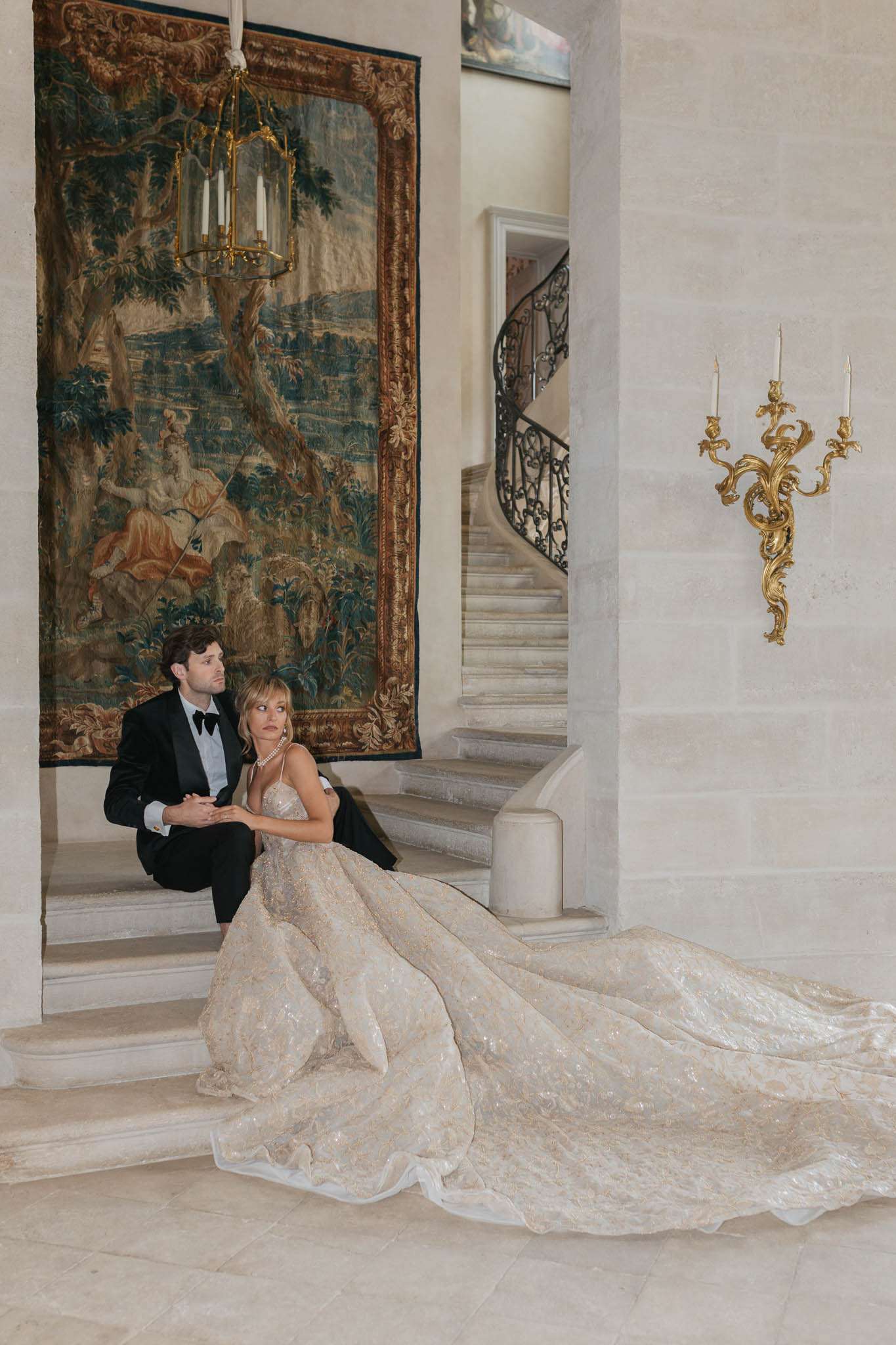A couple portrait taken indoors at what appears to be a French château, with the bride and groom seated on marble steps at the base of a curved wrought-iron staircase. The groom wears a black tuxedo with a bow tie, and the bride wears a heavily embellished ball gown with a champagne and ivory beaded lace bodice and an extremely long, wide cathedral train that spreads across the marble floor. She accessorizes with a pearl or diamond layered necklace. Behind them hangs a large ornate Flemish-style tapestry in rich greens, golds, and earthy tones, framed in gilt, with a gold lantern chandelier suspended above. A gilded rococo-style wall sconce with two candle arms is mounted on the limestone wall to the right. The composition is a full-length portrait shot that deliberately showcases the full spread of the gown's train in the foreground. The overall styling theme is classic French aristocratic. Potential venue feature image.