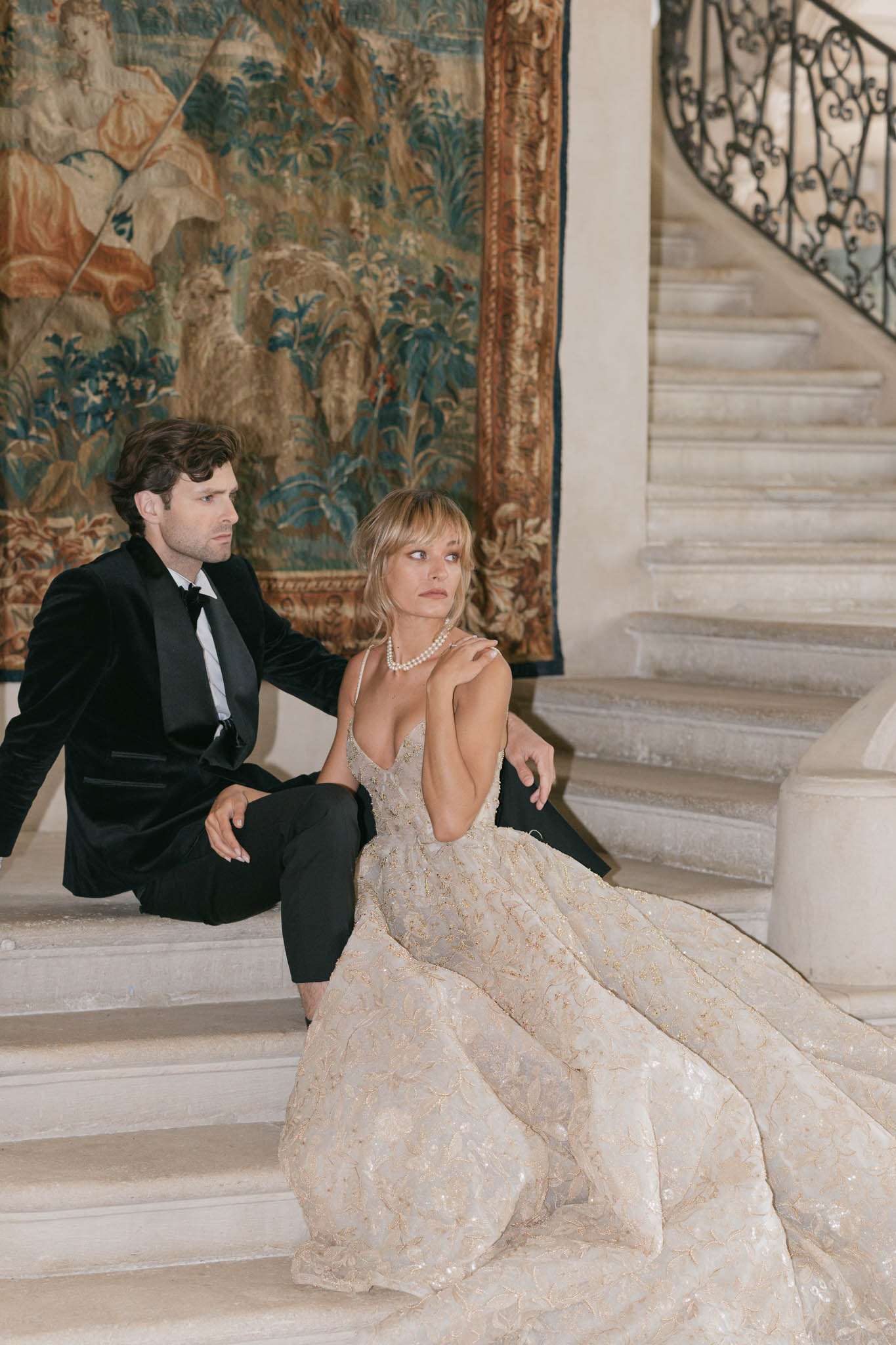 Bride in gold brocade ball gown and groom in black velvet tuxedo seated on curved stone staircase in French chateau
