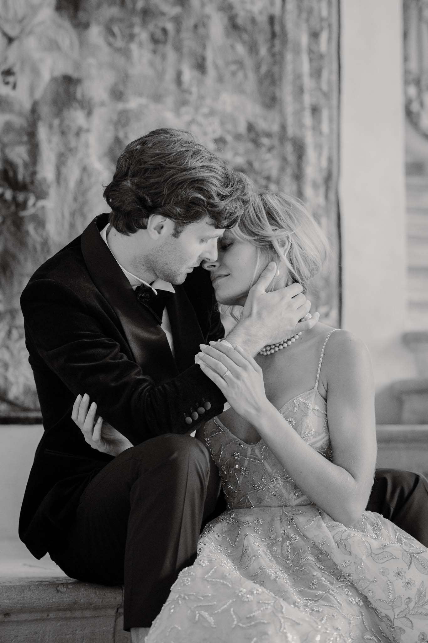 This black-and-white couple portrait shows the bride and groom seated closely together, foreheads nearly touching in an intimate moment. The groom wears a velvet dinner jacket with a bow tie, while the bride wears a heavily embroidered and beaded spaghetti-strap gown with a floral lace pattern and a pearl necklace. His hand cups her face gently as she holds his arm. The background features a large tapestry with dense, painterly tones that suggest an indoor château or historic interior setting. The image is shot in medium close-up with soft contrast and rich dark tones, emphasizing the texture of both the velvet jacket and the embellished dress.