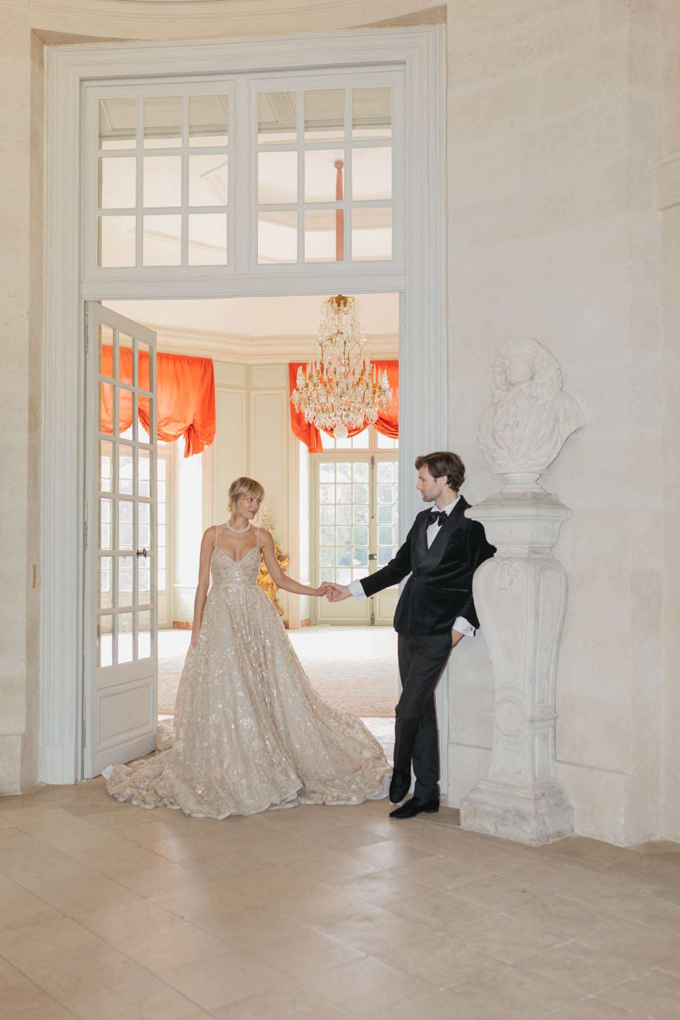 A couple portrait taken indoors at what appears to be a French château, with the bride and groom holding hands through an open set of tall white French doors. The bride wears a heavily embellished champagne-gold ball gown with spaghetti straps, a deep V-neckline, and a full cathedral-length train with floral appliqué detailing at the hem; she accessorizes with a pearl necklace and has her blonde hair in an updo. The groom wears a classic black tuxedo with a bow tie and is leaning casually against a stone pedestal topped with a classical white bust sculpture. Through the open doorway, a gilded crystal chandelier hangs in a paneled room with orange-red draped balloon curtains on tall French windows, giving the space a formal, classical French interior aesthetic. The composition is a full-length portrait shot from a slight distance, framing the couple within the doorway architecture with the chandelier room as a backdrop.