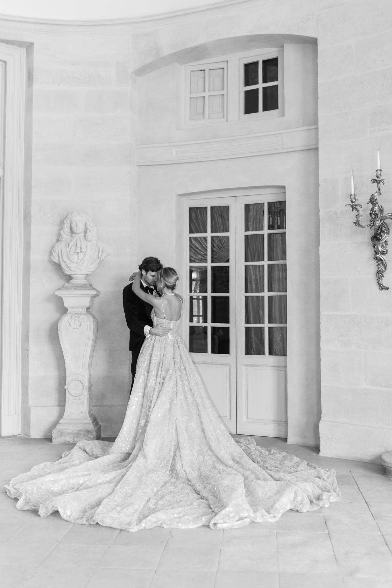 Black-and-white portrait of bride and groom kissing by paneled doors, cathedral train spread across stone floor