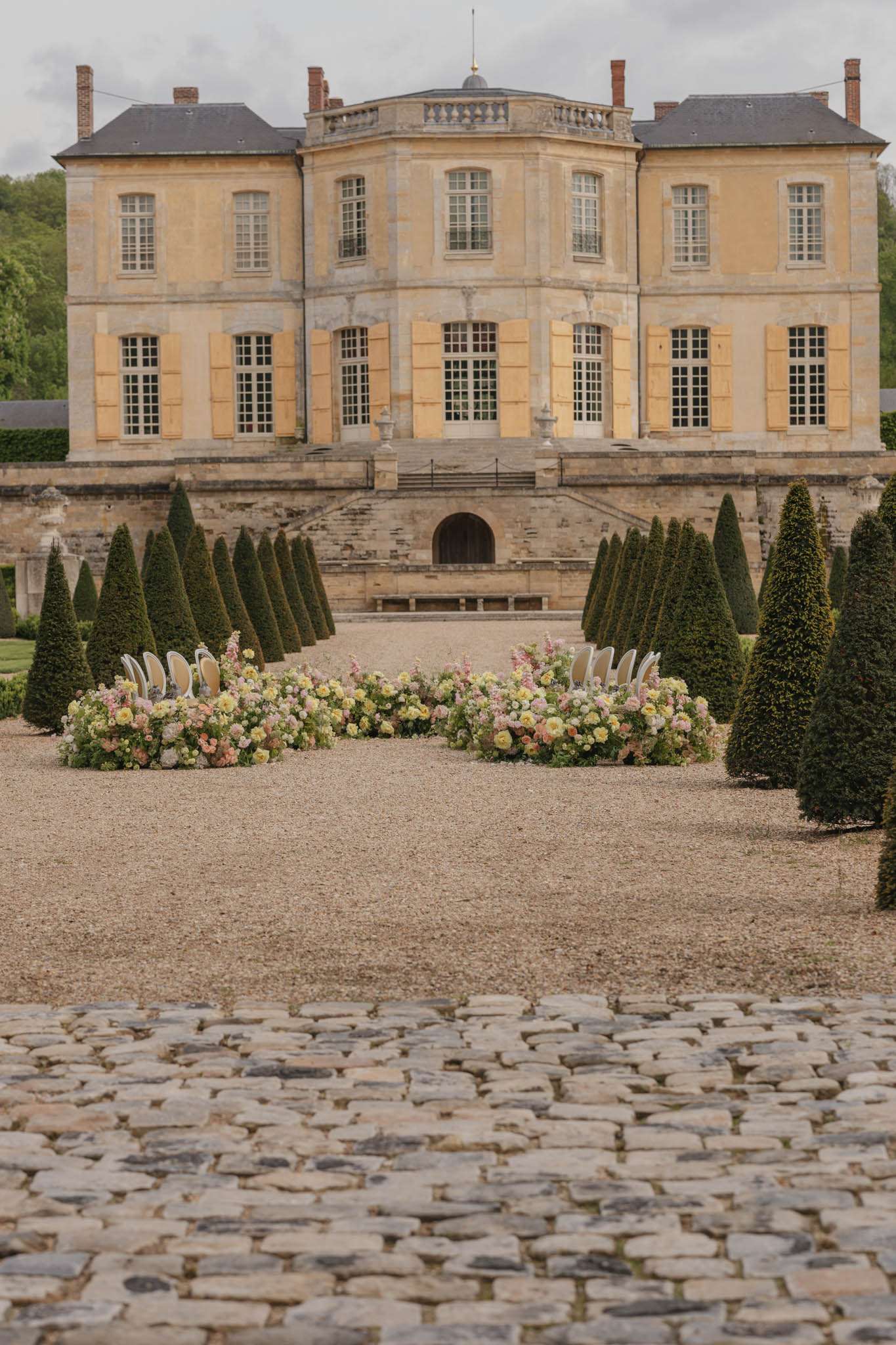 An outdoor wedding ceremony setup photographed from a wide, elevated angle in the formal garden of a French château. The ceremony area features two curved rows of white chairs flanking a central aisle, bordered by large ground-level floral installations in soft pink, blush, yellow, and peach tones — appearing to include garden roses, delphiniums, and ranunculus arranged in lush, low mounds. The formal garden is lined with precisely clipped conical topiary trees leading toward the château facade, which is a three-story honey-colored limestone building with yellow shutters, arched windows, and a slate mansard roof. The foreground shows a cobblestone forecourt, and the overall styling is classic French formal with a soft, romantic floral palette. Potential venue feature image.
