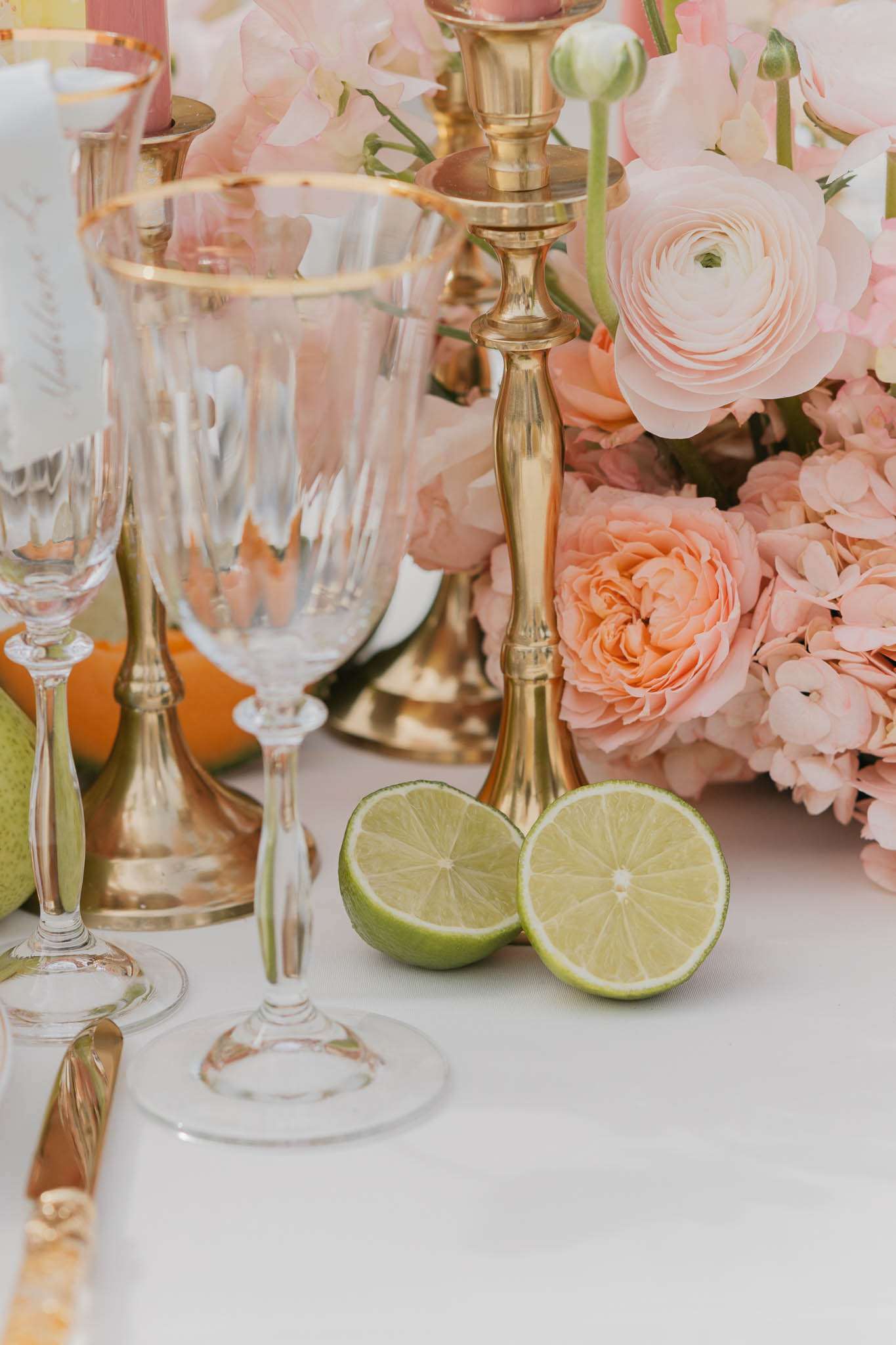 A close-up detail shot of a wedding reception tablescape styled with a peach, blush, and gold palette. The floral centerpiece includes blush ranunculus, peach garden roses, and pink hydrangeas, arranged alongside tall brass candlesticks holding pale pink taper candles. Crystal stemware with gold-rimmed detailing and a gold-handled knife are placed on a white linen tablecloth, with a halved lime and green pears used as decorative fruit accents alongside what appears to be an orange. The overall styling mixes classic and garden-inspired elements with a warm, fruit-forward tablescape concept.