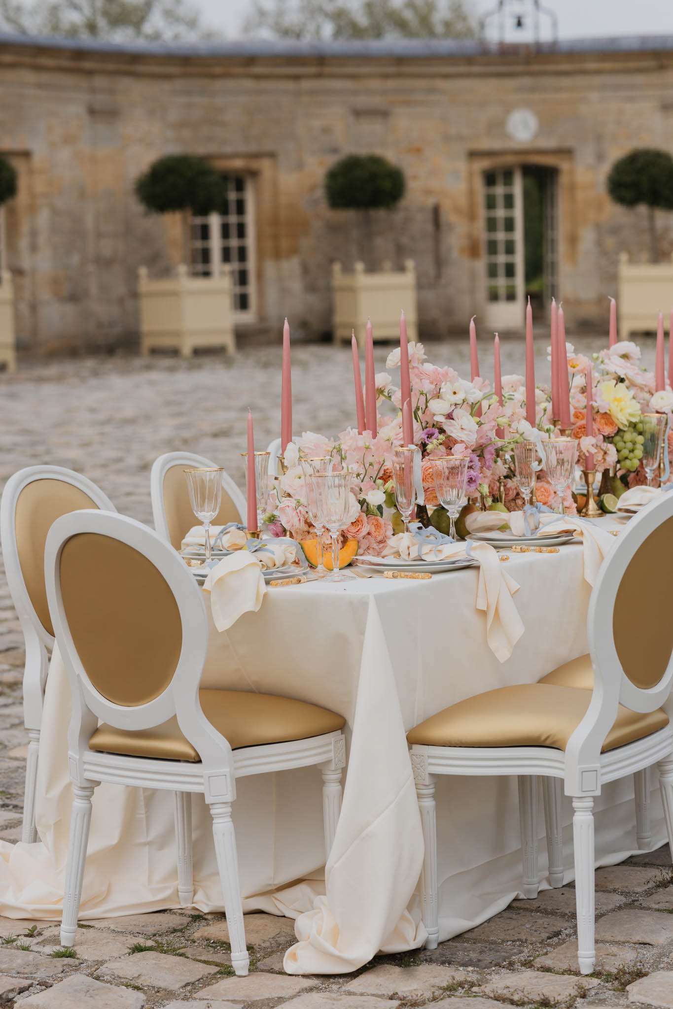 Long reception table on chateau courtyard with blush roses peach ranunculus dusty rose candles and gold-rimmed crystal