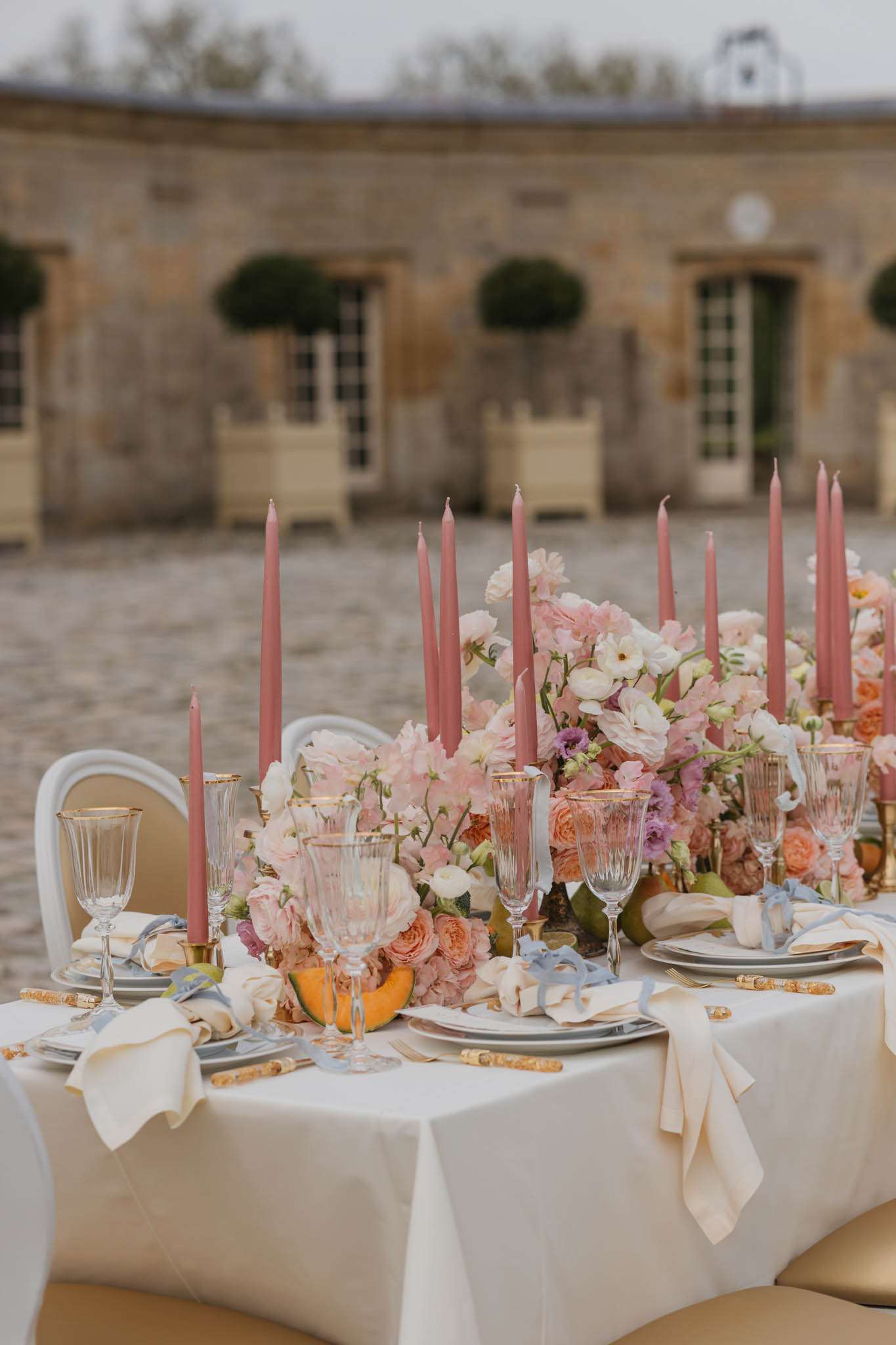A close-up detail shot of an outdoor reception tablescape set in the cobblestone courtyard of a classic French château, visible in the background. The white linen-covered table features a lush floral runner composed of blush pink garden roses, peach ranunculus, white sweet peas, mauve hydrangeas, and small purple blooms, interspersed with whole melons and pears as decorative fruit elements. Tall dusty rose taper candles in gold candlestick holders rise from within the floral arrangement across the length of the table. Place settings include charcoal grey rimmed plates layered on ivory chargers, crystal glassware with gold-rimmed detailing, gold-handled flatware, and ivory linen napkins tied with pale blue ribbon. The overall decor palette combines dusty rose, peach, ivory, soft grey, and muted gold in a refined classic French style.