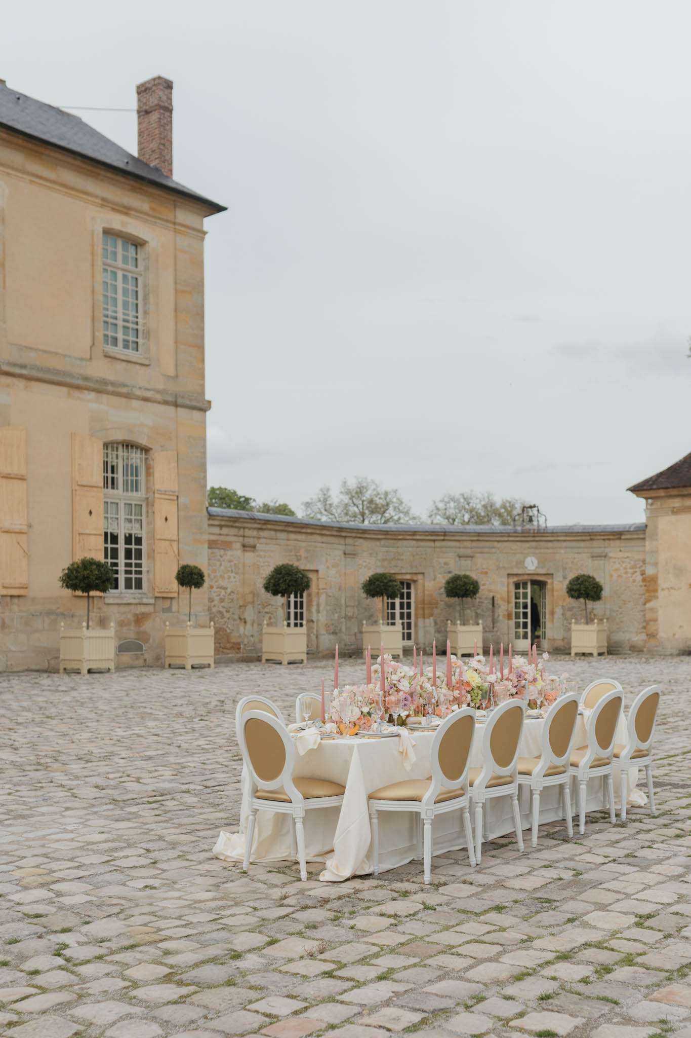 Long reception table with floral runner and pink candles set in chateau courtyard with stone colonnade behind