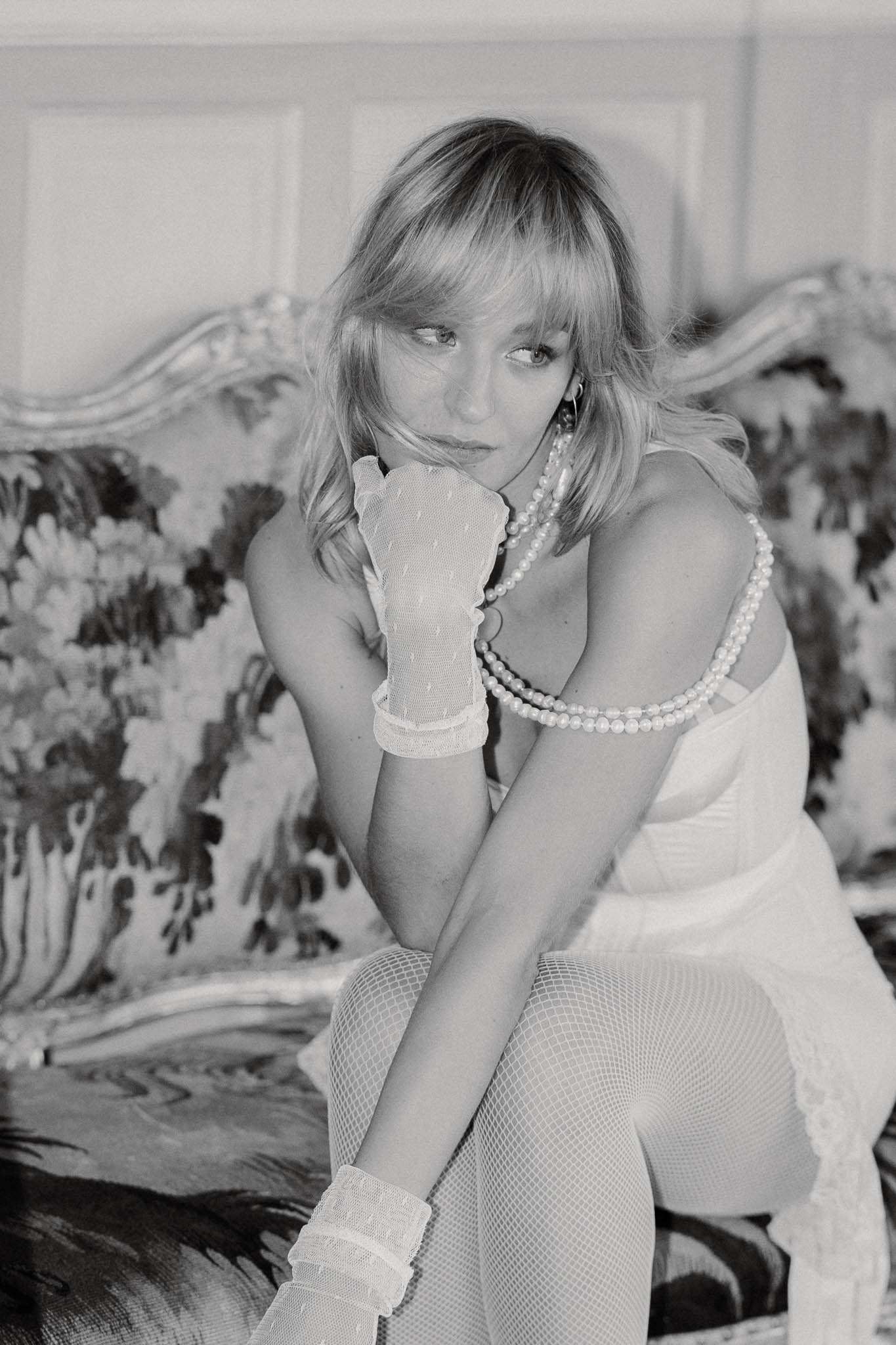 A black-and-white bridal getting-ready portrait of a single woman seated on an ornate floral-upholstered sofa with carved detailing, set against a paneled interior wall. She wears a white corset-style top, white fishnet tights, short lace-trimmed gloves, and layered pearl necklaces, with hoop earrings and loose blonde hair with a fringe. Her pose is relaxed and slightly turned, with one gloved hand raised to her chin. The styling has a retro-inspired bridal aesthetic combining pearl accessories with edgy fishnet and lace details. The image is a close-to-medium portrait shot with soft contrast and light grain typical of film photography.