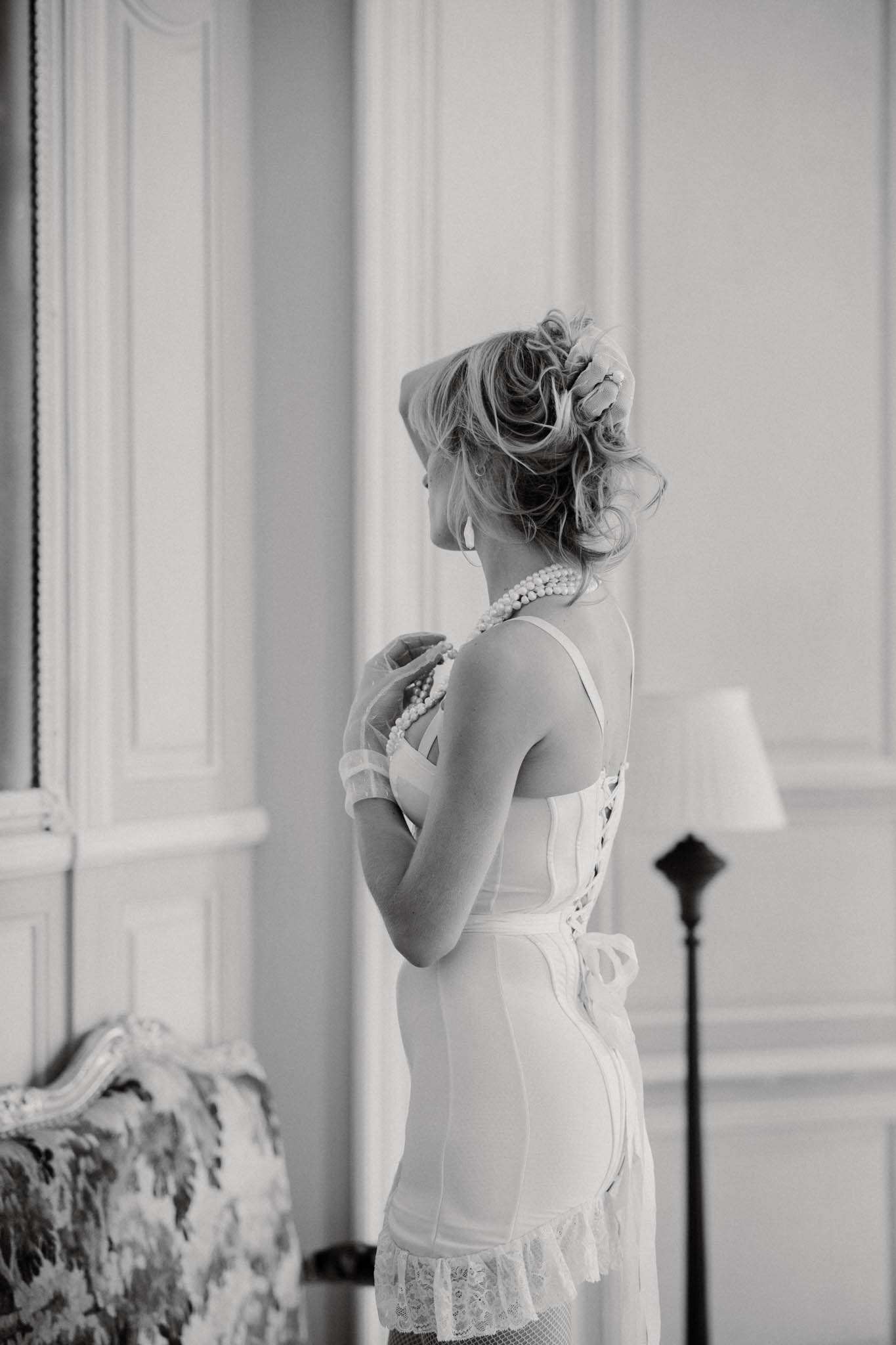 Black and white portrait of bride in bridal slip fastening pearl necklace in chateau interior