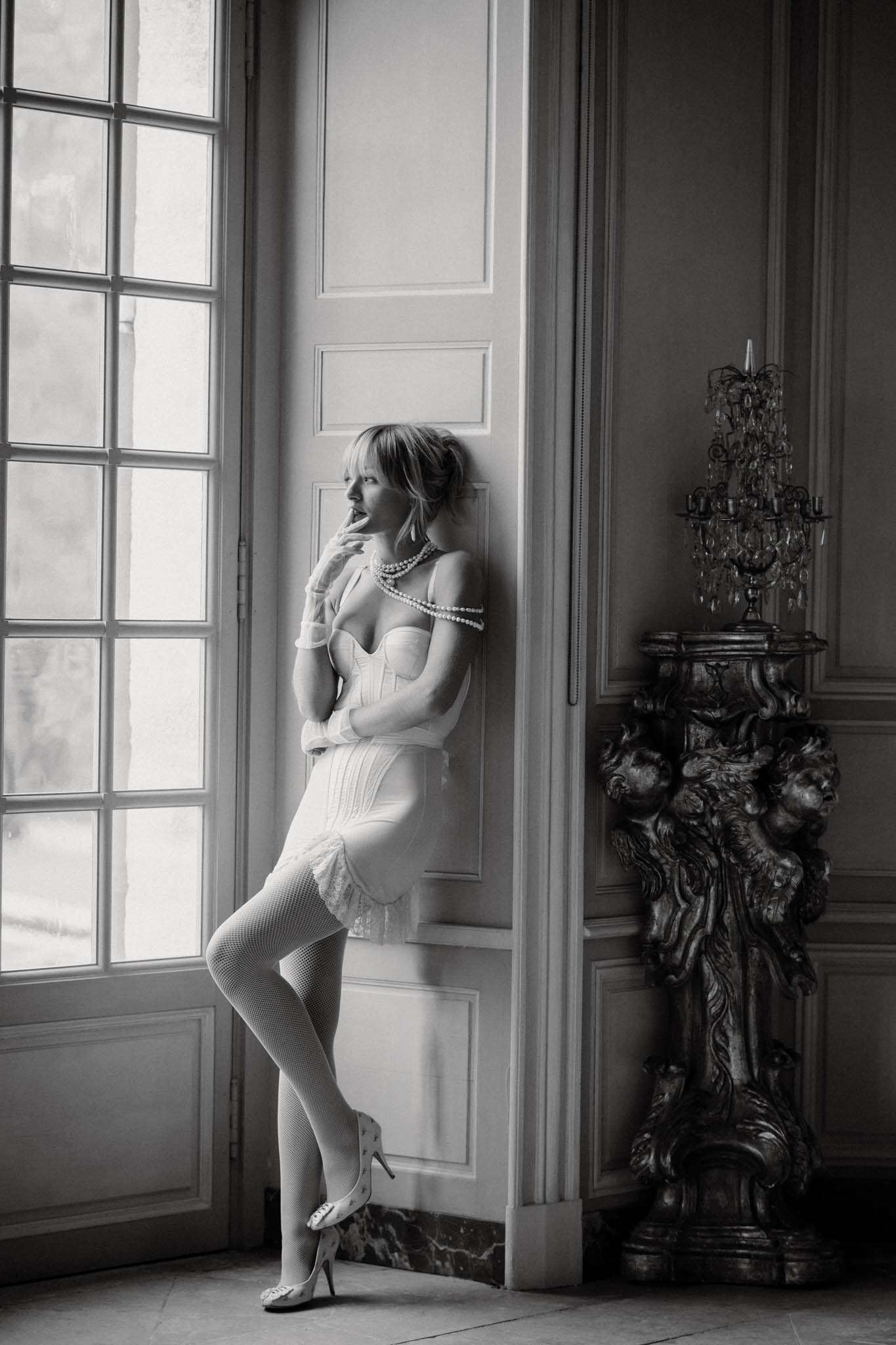 Black-and-white bridal boudoir portrait taken indoors in what appears to be a French château or manor house. The bride leans against a panelled door between tall French windows, wearing a structured corset-style bridal bodysuit with lace shorts, fishnet stockings, white heeled mules, layered pearl necklaces, and short white gloves. Her hair is styled in a loosely pinned updo with a fringe, and she gazes toward the window with one hand raised to her lips. To her right stands an ornate carved pedestal topped with a crystal candelabra. The image has soft, even tones with moderate contrast, and is composed as a full-length portrait with the architectural detailing of the room forming the background.