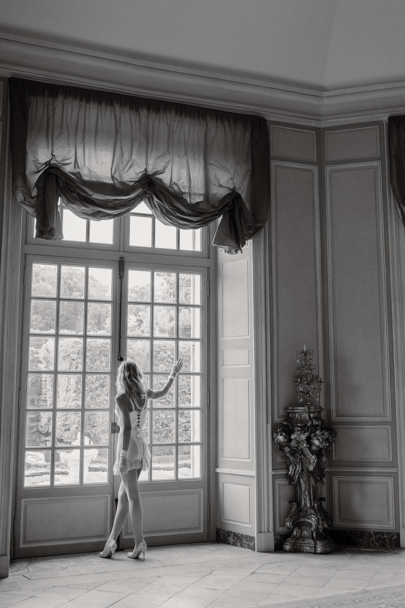 This black-and-white image captures a bride during a getting-ready moment, standing alone in a grand interior room and reaching toward a set of tall French doors. She is wearing a corset-style bridal bodysuit with lace trim, suspenders, stockings, and heeled shoes, with a long pearl necklace visible at her back. The room features classic French boiserie wall paneling, a marble-tiled floor, draped swag curtains over the tall windows, and an ornate gilt candelabra or torchère on a marble plinth to the right. The composition is a full-length portrait shot from behind, with strong backlighting from the window creating high contrast between the bright exterior and the mid-tone interior walls.