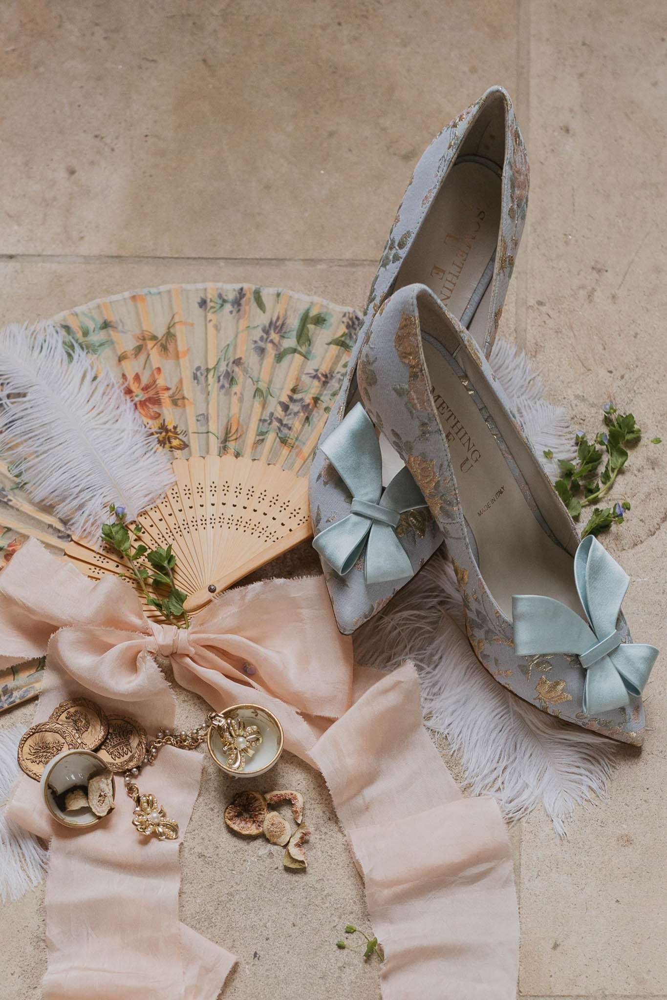 A flat lay detail shot of bridal accessories arranged on a stone floor. The central items are a pair of pale blue brocade pointed-toe heels with gold floral embroidery and mint satin bow embellishments, labeled 'Something Bleu' and made in Italy. Surrounding them are two hand fans — one painted with a floral motif on bamboo slats, one plain perforated wood — along with white feather trim, a large blush pink silk ribbon bow, gold coin jewellery pieces, a delicate gold floral earring or charm with a pearl detail, a small circular gold compact or trinket, and scattered small botanical sprigs. The overall styling palette is soft blush, mint, and gold, with a romantic, vintage-inspired aesthetic.
