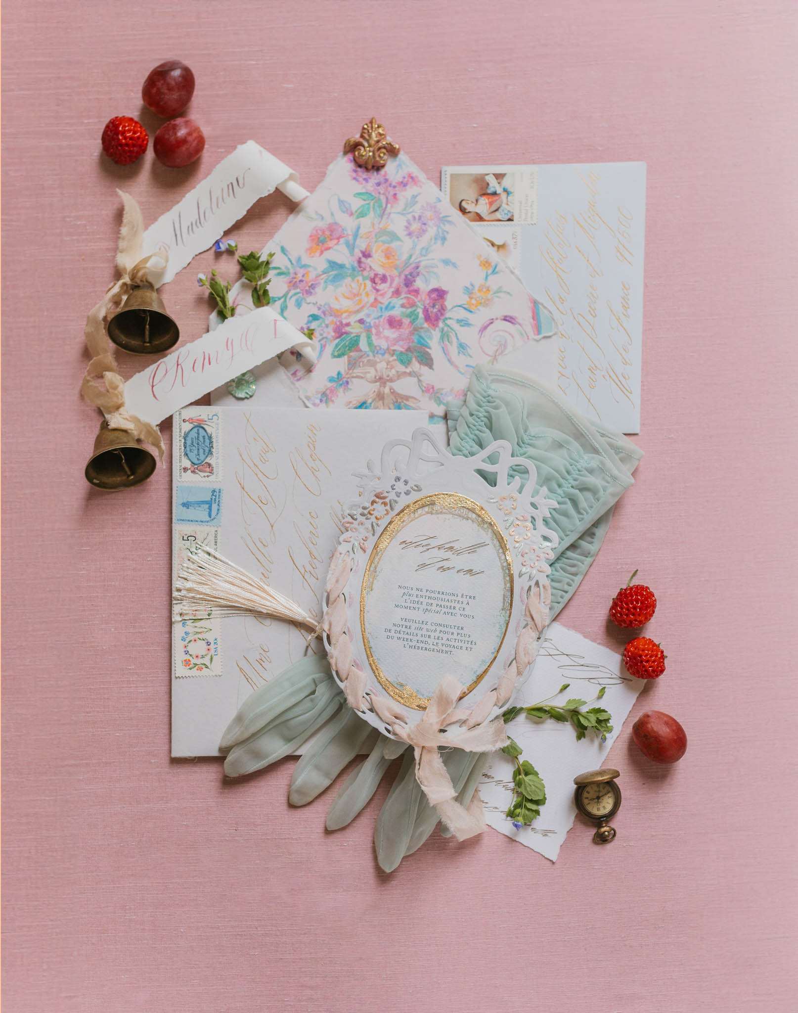 A flat lay styled detail shot of a French wedding stationery suite arranged on a dusty pink linen surface. The collection includes multiple envelopes and cards featuring gold calligraphy script, a watercolor floral-lined envelope with a decorative gold fleur-de-lis seal, a sage green envelope with vintage postage stamps, a pale blue card with gold handwritten text, and a central invitation card with a gold oval frame set within white laser-cut lace detailing, tied with a blush silk ribbon and accompanied by sage green feathers. Styling props include small brass bells tied with cream ribbon, a miniature brass pocket watch, fresh red strawberries and small dark plums, and scattered herb sprigs. The overall palette combines dusty pink, sage green, ivory, gold, and soft blue, with a romantic vintage French aesthetic.
