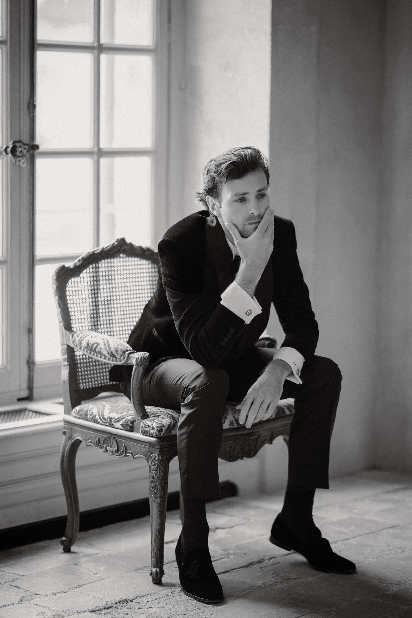 Black and white groom in dark suit seated in Louis XV caned armchair beside French windows