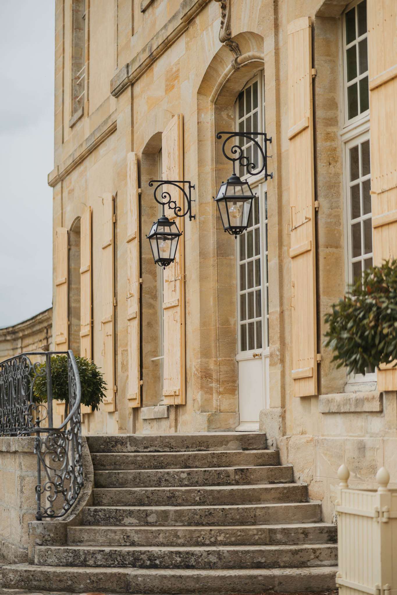 Honey-toned limestone chateau facade with stone staircase, wrought-iron lanterns, and trimmed topiaries