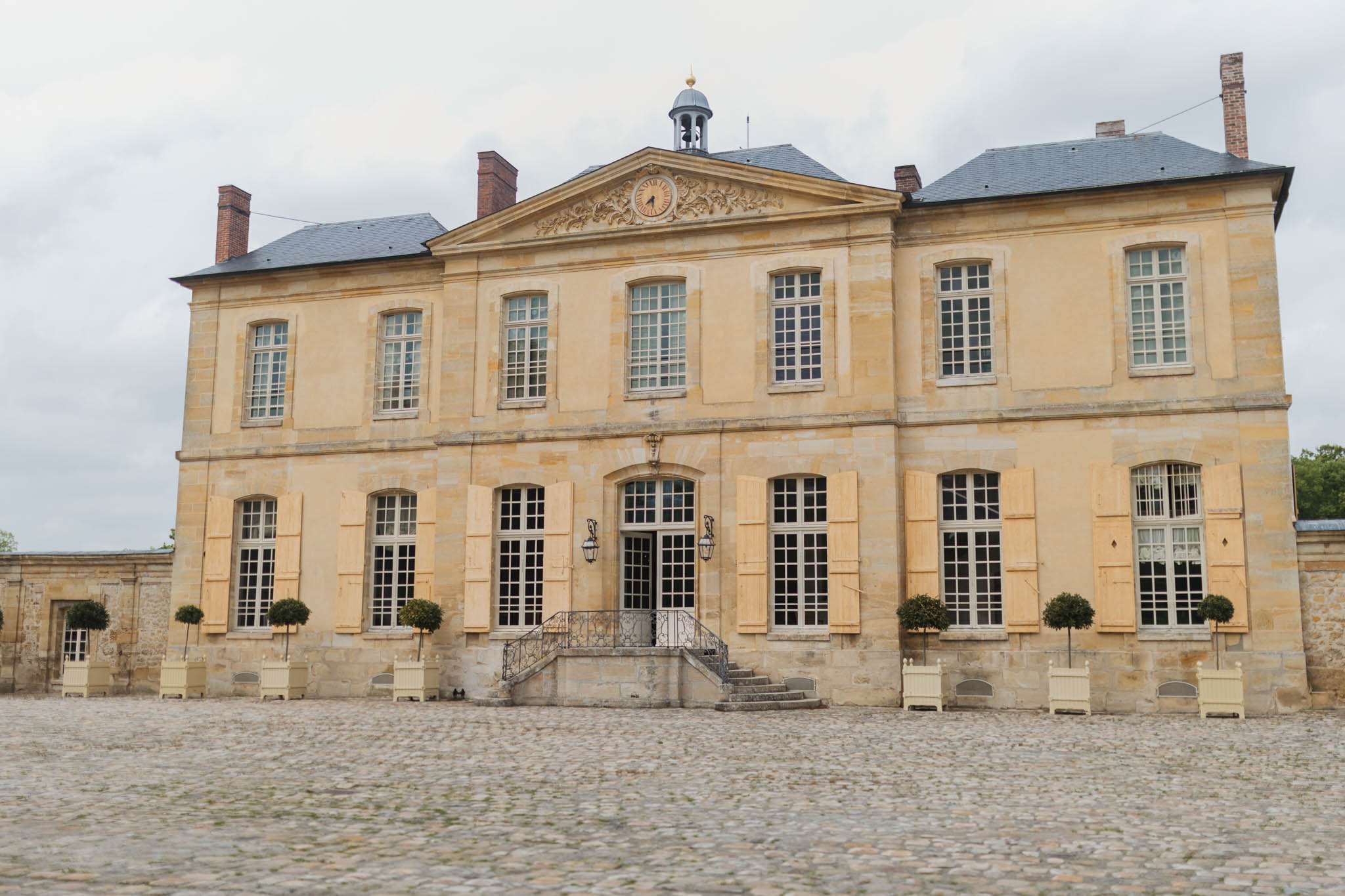 Wide shot of a French château facade featuring a two-story neoclassical building constructed in honey-colored limestone with slate grey mansard-style roofing and multiple brick chimneys. The central entrance is framed by a decorative pediment with a carved clock and gold finial cupola above, flanked by tall arched windows with cream-painted shutters and iron lanterns on either side of the door. A symmetrical arrangement of clipped topiary trees in cream wooden planters lines the front of the building, and a cobblestone courtyard extends across the foreground. No people are present in the image. Potential venue feature image.