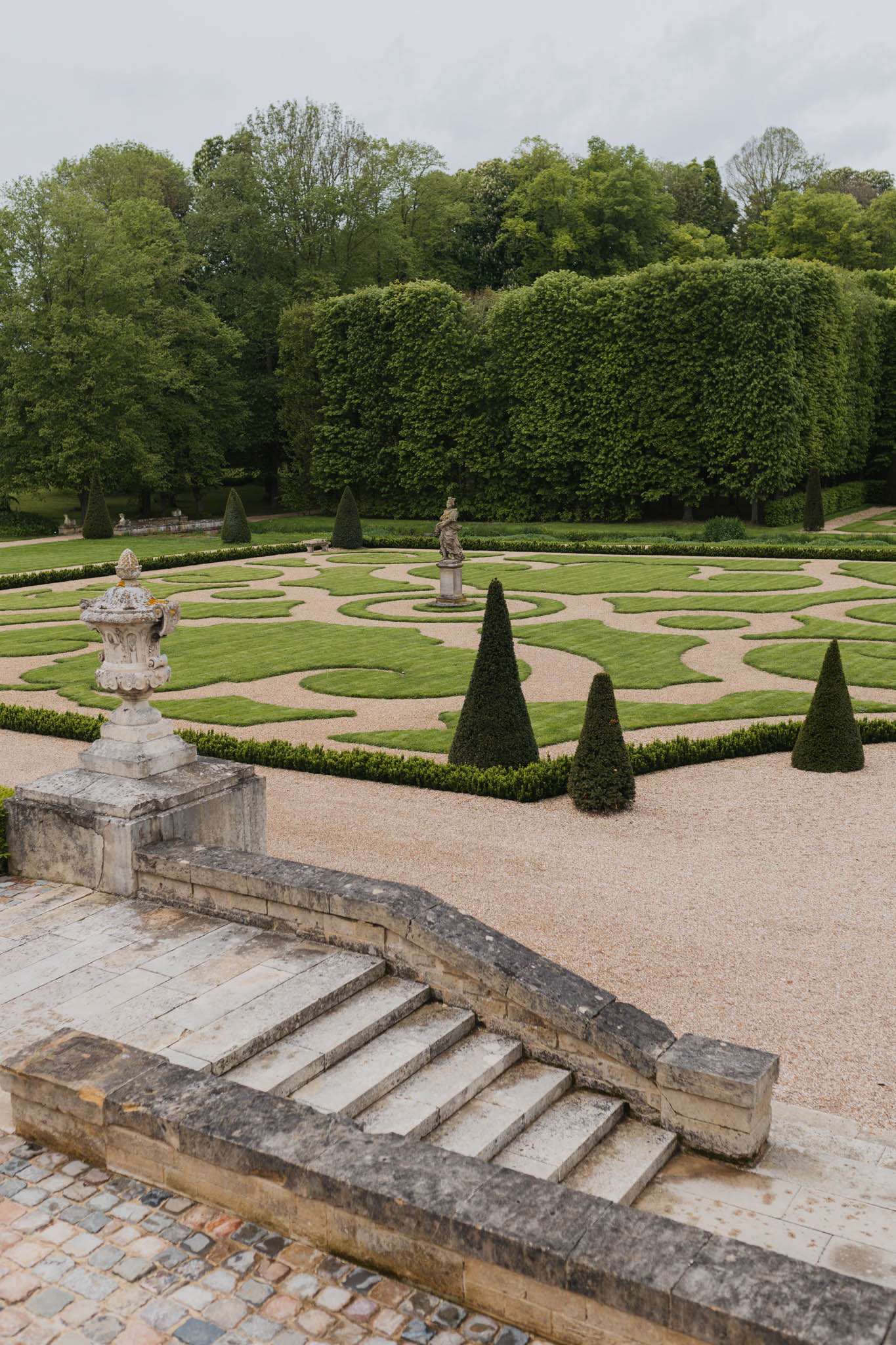 This image shows no people and captures the formal French-style gardens of a château venue, shot from an elevated vantage point overlooking a wide exterior staircase with stone balustrades and a decorative stone urn. The garden below features a classic parterre design with low box hedging clipped into scrolling geometric patterns set within a gravel surround, cone-shaped topiary yews, a central stone statue on a pedestal, and a backdrop of densely clipped hornbeam hedges. The foreground includes worn stone steps with cobblestone paving, suggesting the image is taken from a terrace or upper landing. Wide establishing shot taken outdoors under overcast light. Potential venue feature image.
