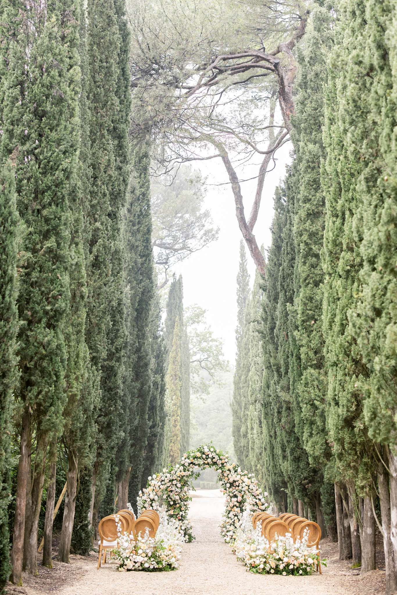 Circular rose arch in blush and peach on cypress-lined gravel allee with chiavari chairs on each side