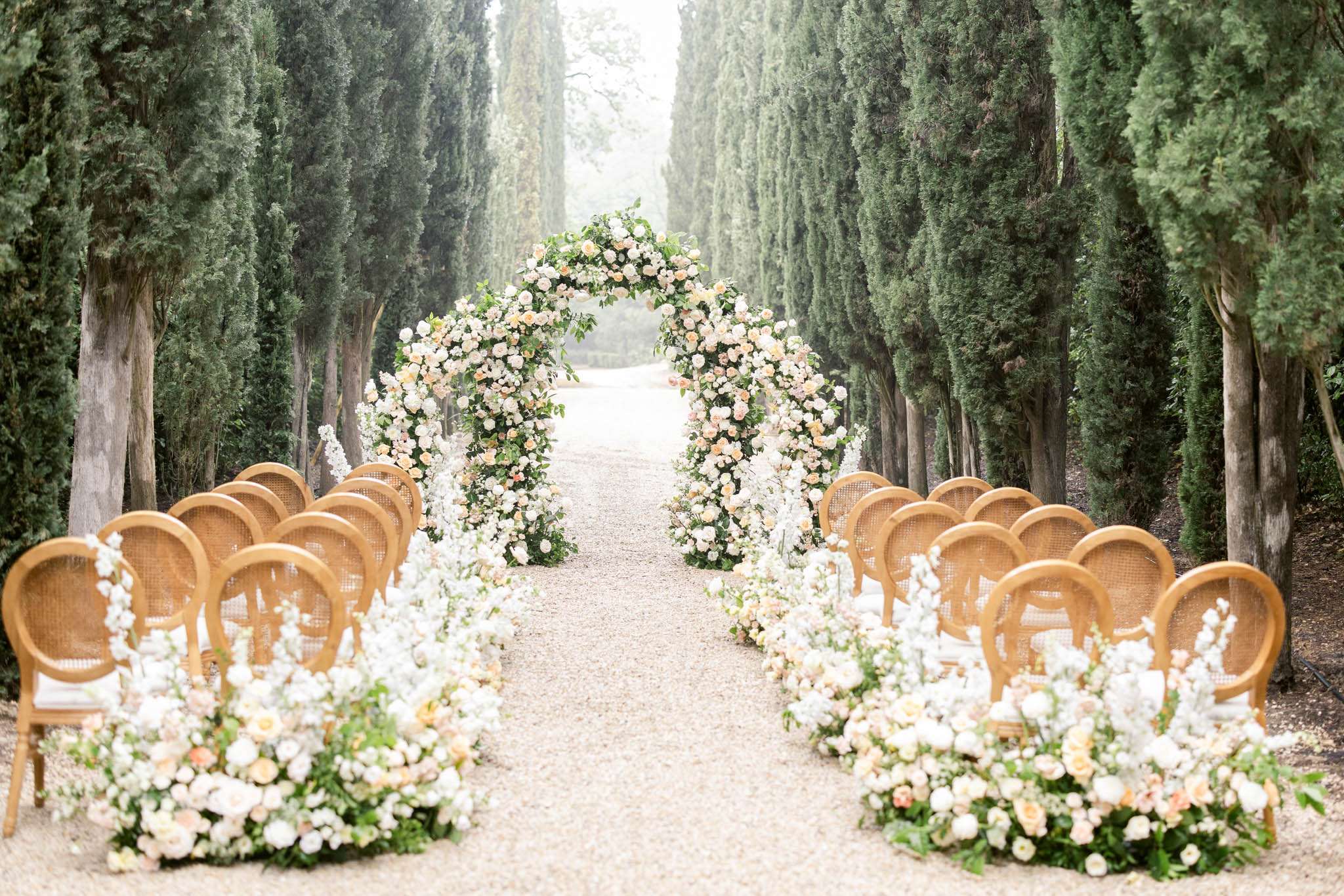 An outdoor ceremony setup on a gravel allée lined with tall cypress trees, photographed before the ceremony with no guests or couple present. A large circular floral arch serves as the focal point at the end of the aisle, densely covered in white roses, peach garden roses, white delphinium, and green foliage. The aisle is flanked on both sides by rows of natural wood cane-back Louis XVI-style chairs, with lush low floral arrangements at the base of each row featuring the same white and peach rose palette with trailing greenery. The color palette is soft white, cream, and peach throughout all floral elements. The composition is a wide symmetrical shot taken from the guest entry point looking toward the arch, creating a strong central perspective. Classic French style with a romantic garden aesthetic. Potential venue feature image.