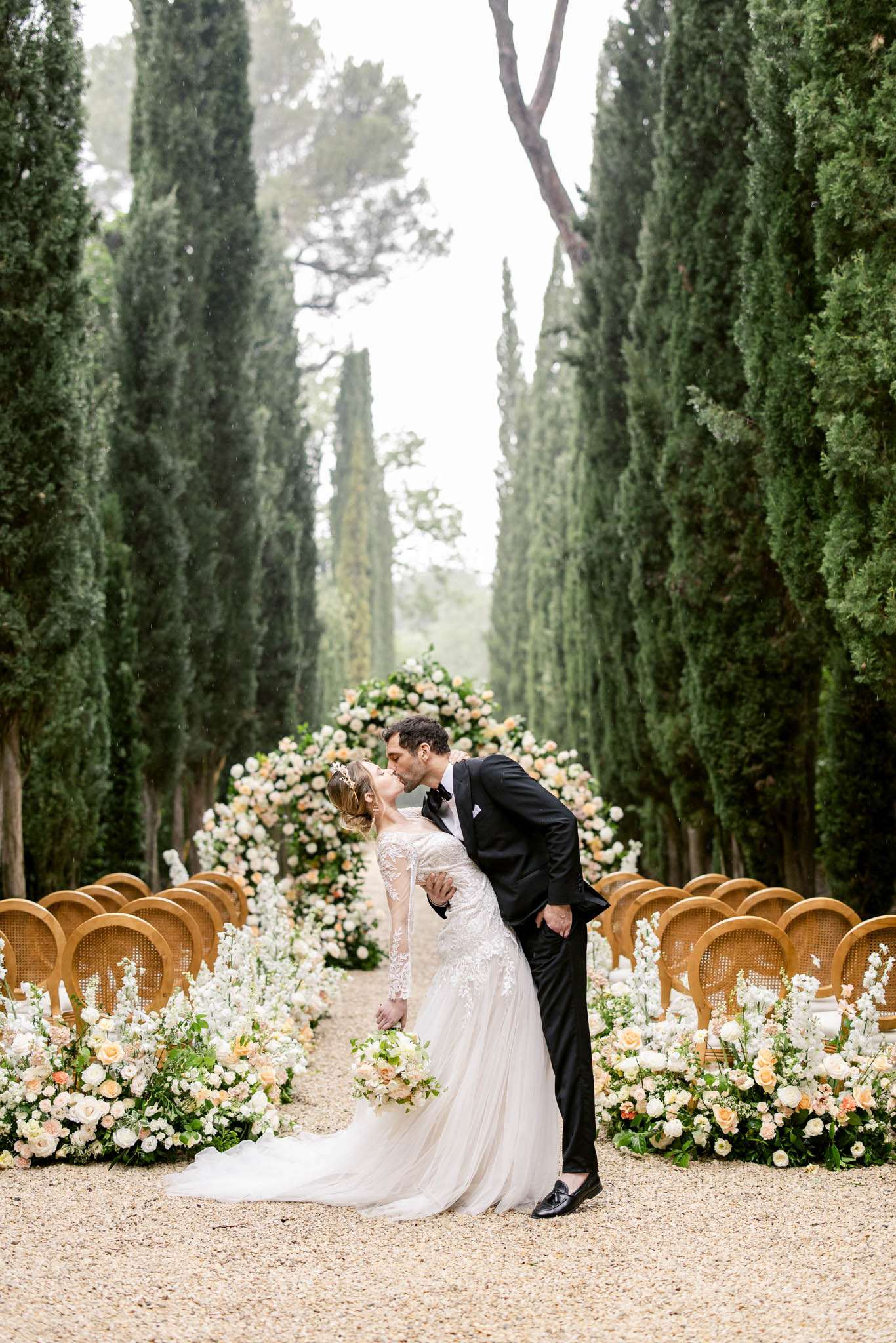 Bride and groom dip kiss under circular floral arch on cypress-lined gravel aisle with cane-back chairs