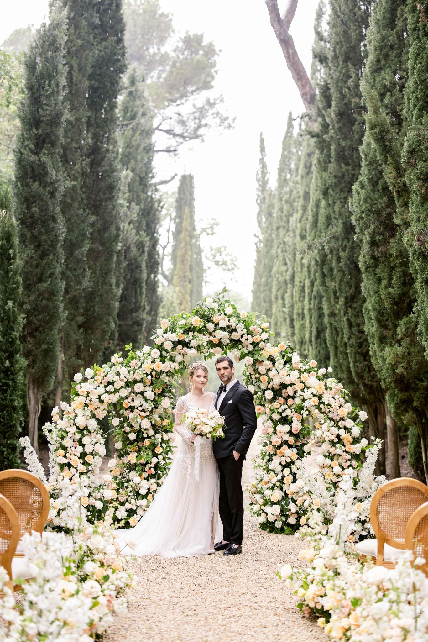 Bride and groom at circular rose arch on cypress-lined allee with aisle floral arrangements