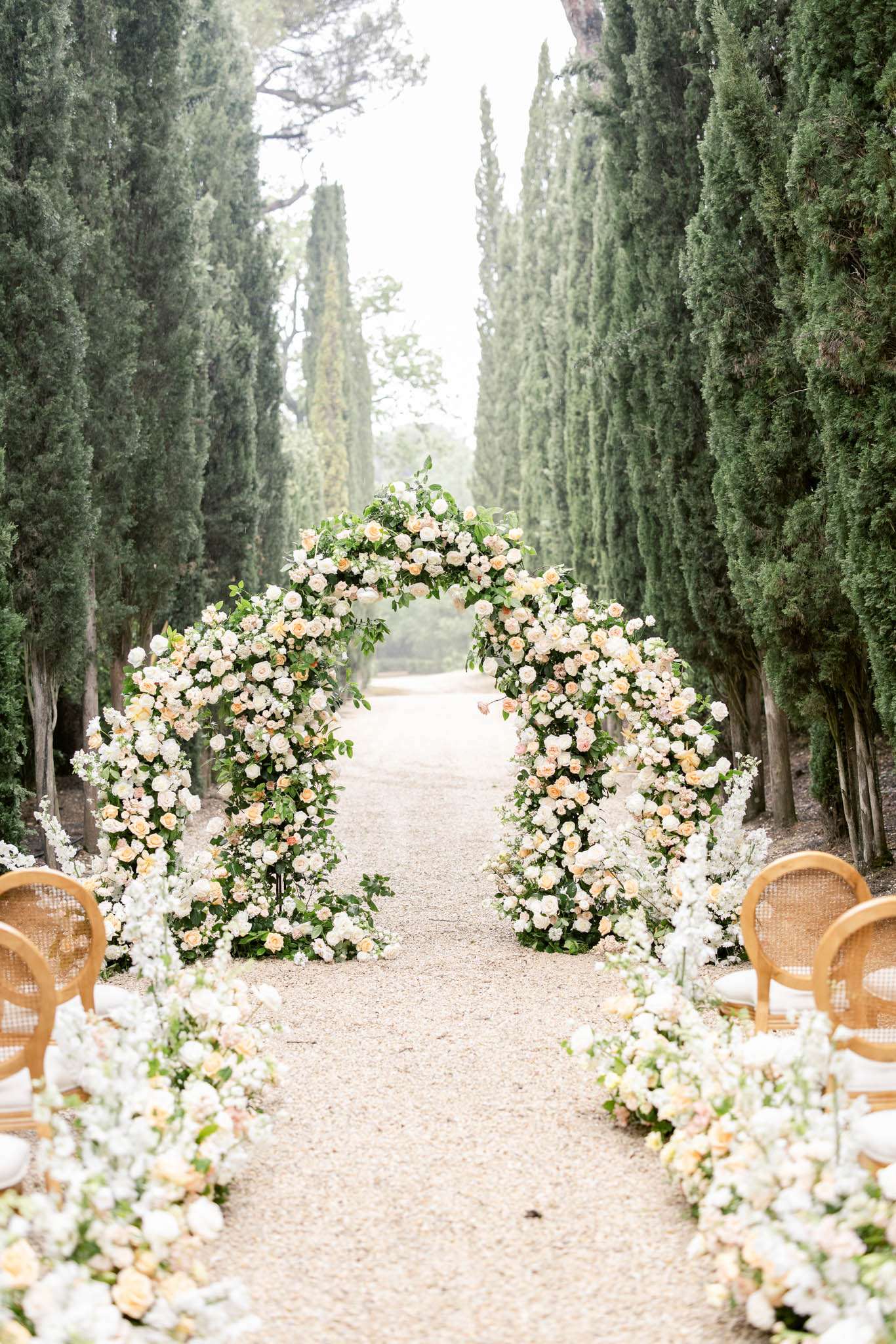 Outdoor ceremony aisle lined with cypress trees and two circular floral arches in white and peach roses with cane-back chairs