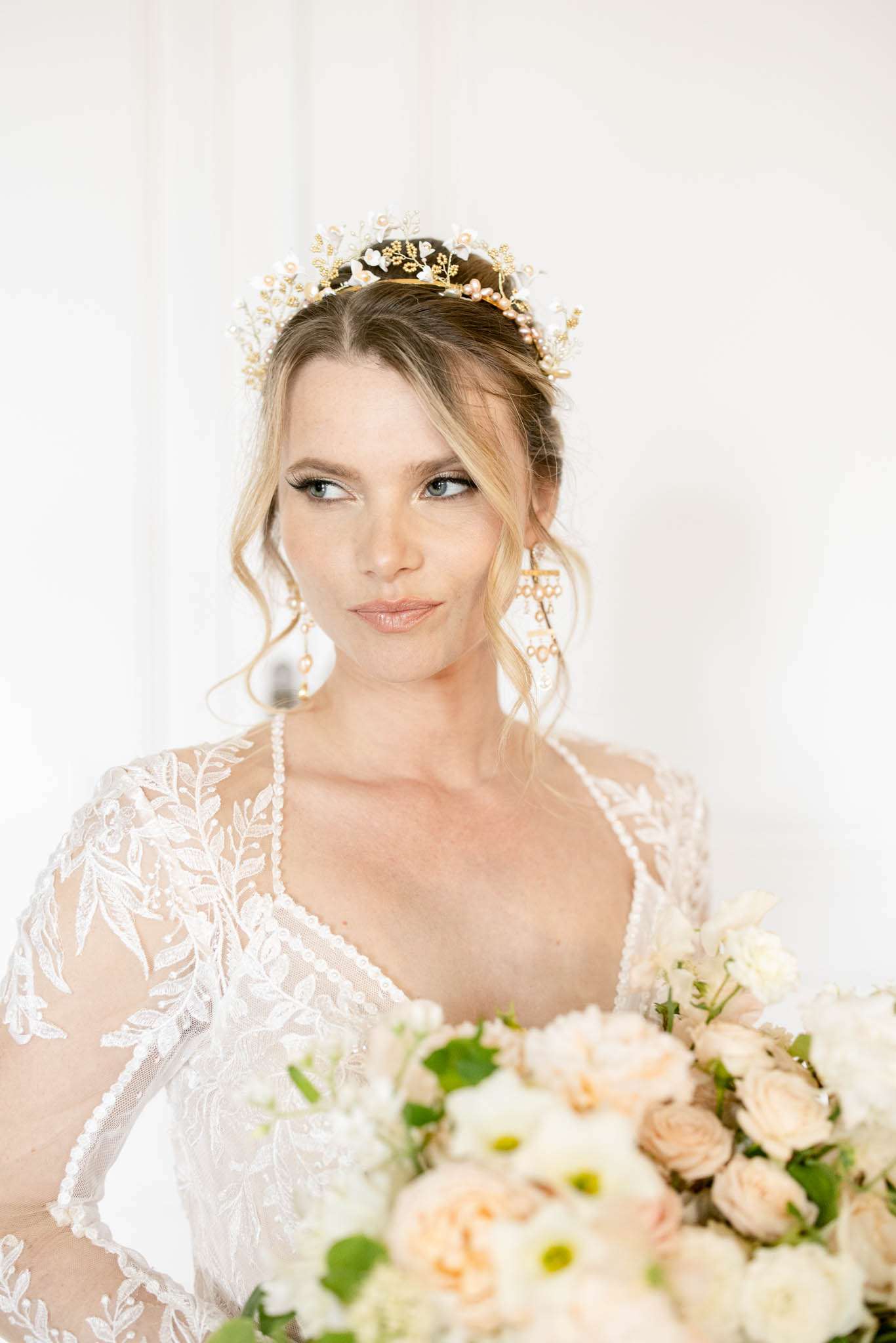 Bride in white lace dress with sheer sleeves holding blush rose bouquet, wearing gold floral crown and pearl earrings