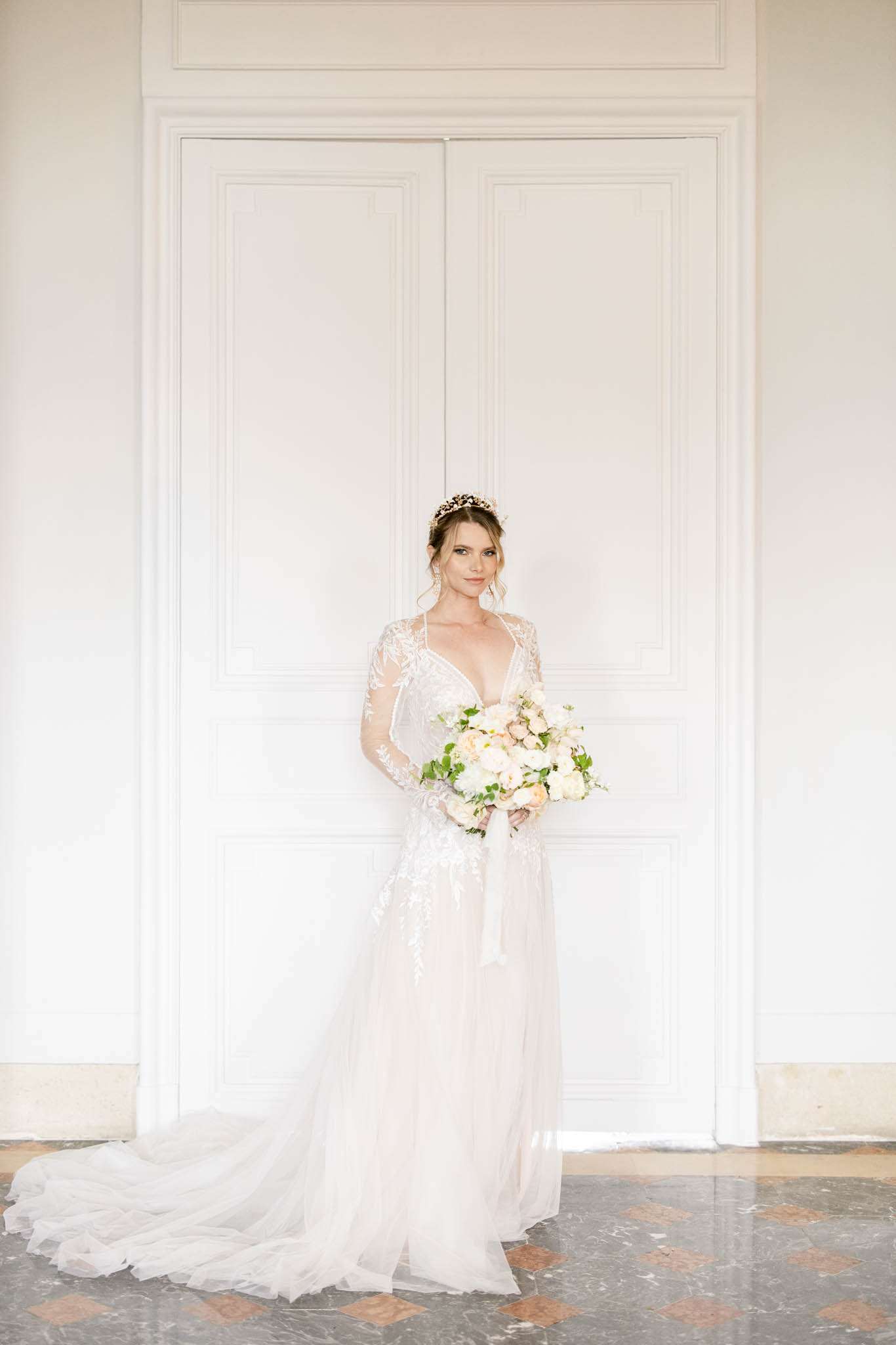 A full-length bridal portrait taken indoors against a white panelled double door, likely inside a château or classical French venue with marble-tiled flooring visible at the base. The bride wears an ivory A-line gown with sheer, lace-embroidered long sleeves, a deep V-neckline, and a floor-length tulle train. She accessorises with a gold floral crown and drop earrings, with her hair styled in a soft updo. She holds a loose, generously sized bouquet of white and blush garden roses, peonies, and soft greenery tied with an ivory ribbon. The overall styling aesthetic is classic and formal, consistent with a French heritage venue setting.